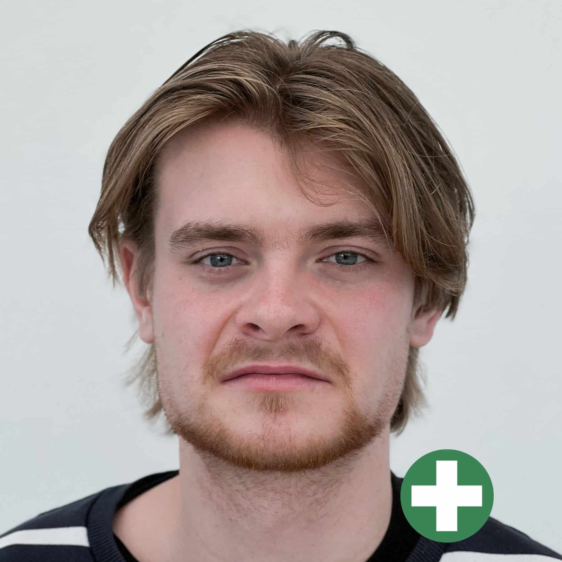 A young man with light brown hair and a short beard looks at the camera with a neutral expression. He is wearing a black and white striped shirt. A green circle with a white plus sign is overlaid in the bottom right corner.