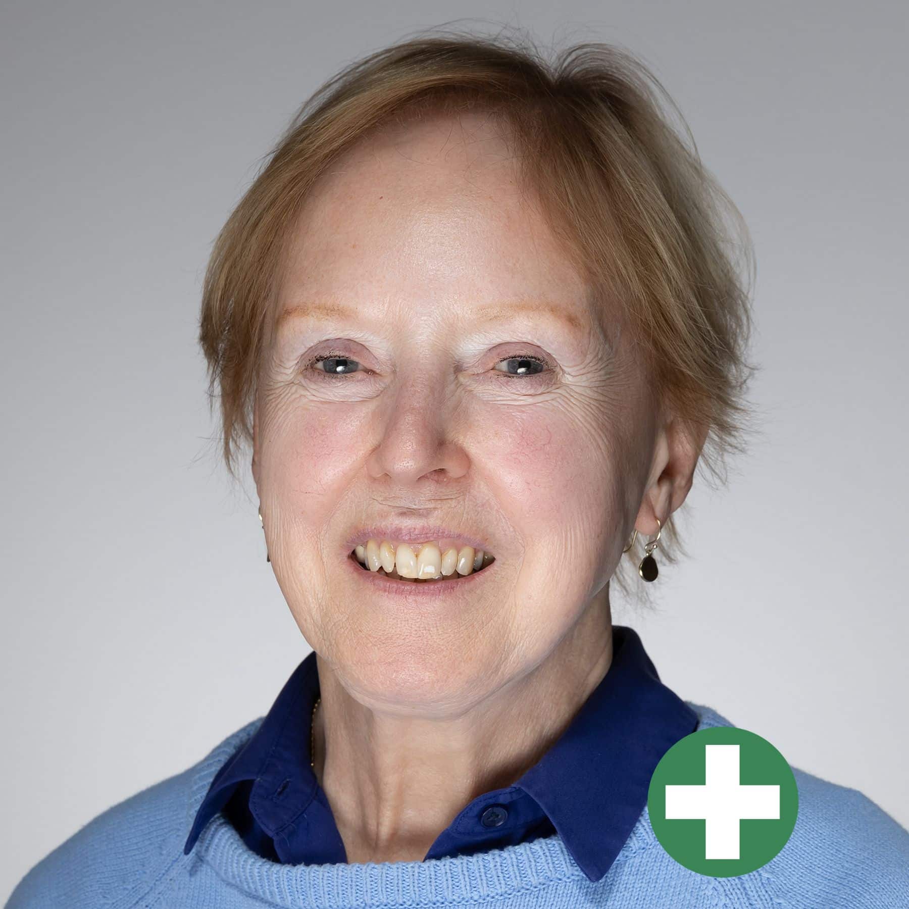 Smiling older woman with short light brown hair, wearing a blue jumper and navy collared shirt. A green circle with a white plus symbol is overlaid in the bottom right corner.