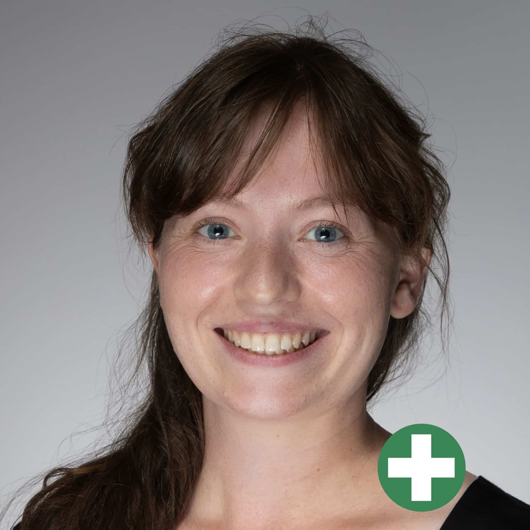 A smiling woman with long brown hair and blue eyes, wearing a black top, is shown against a light grey background. A green circle with a white plus sign is in the lower-right corner of the image.
