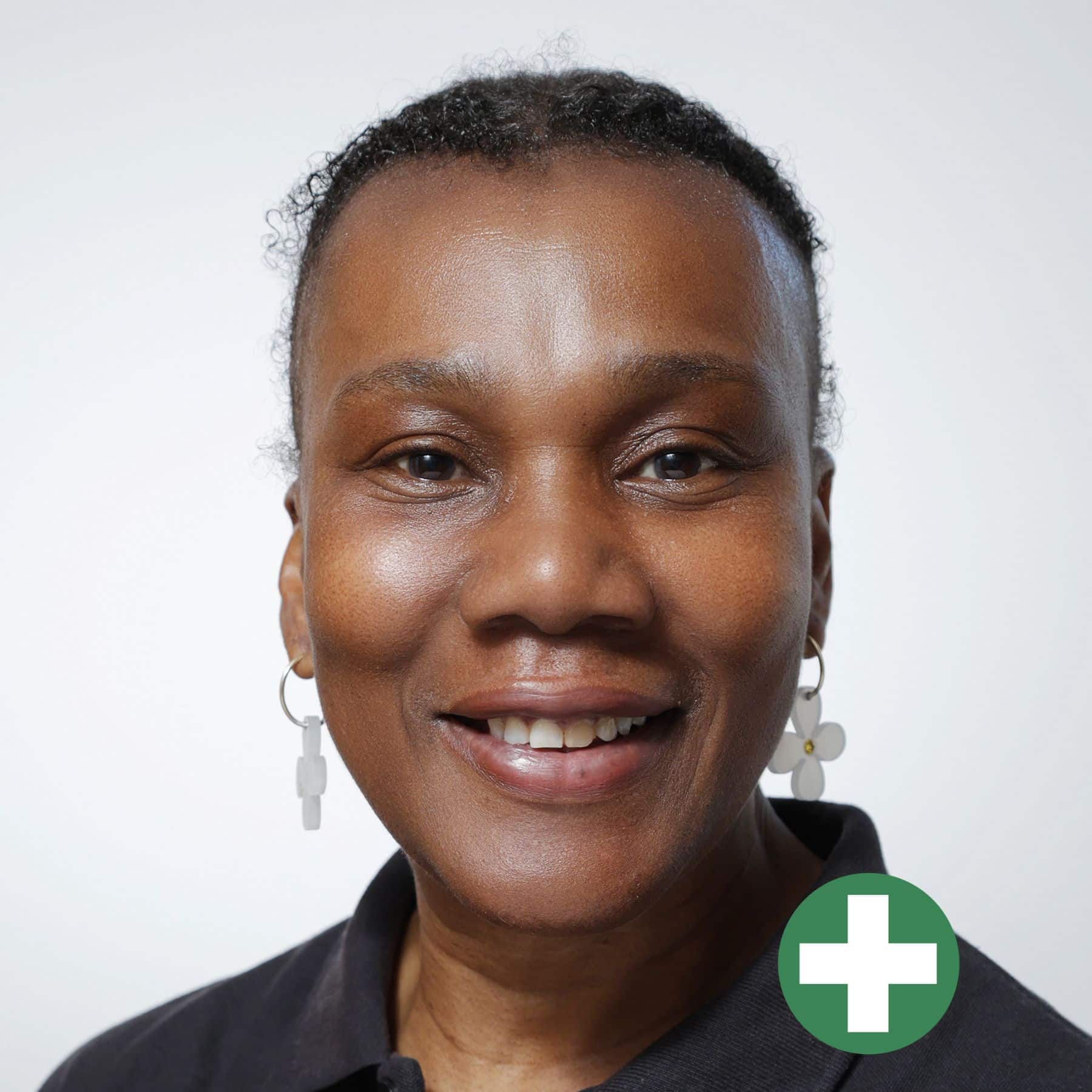 A smiling woman with short curly hair and dangling earrings, wearing a dark collared shirt, is shown against a plain light background. A green circle with a white plus sign is overlaid in the lower right corner.