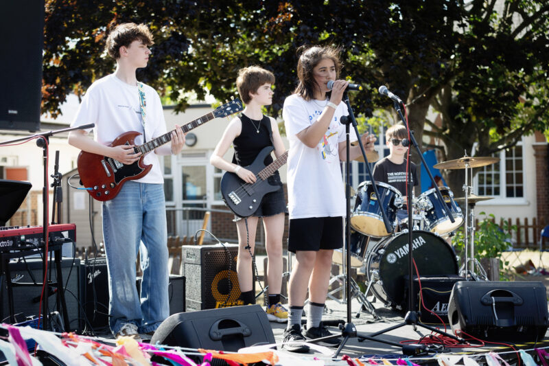 A four-person band performs outdoors. The guitarist, bassist, singer, and drummer are playing on a stage decorated with colourful streamers, with trees and buildings in the background.