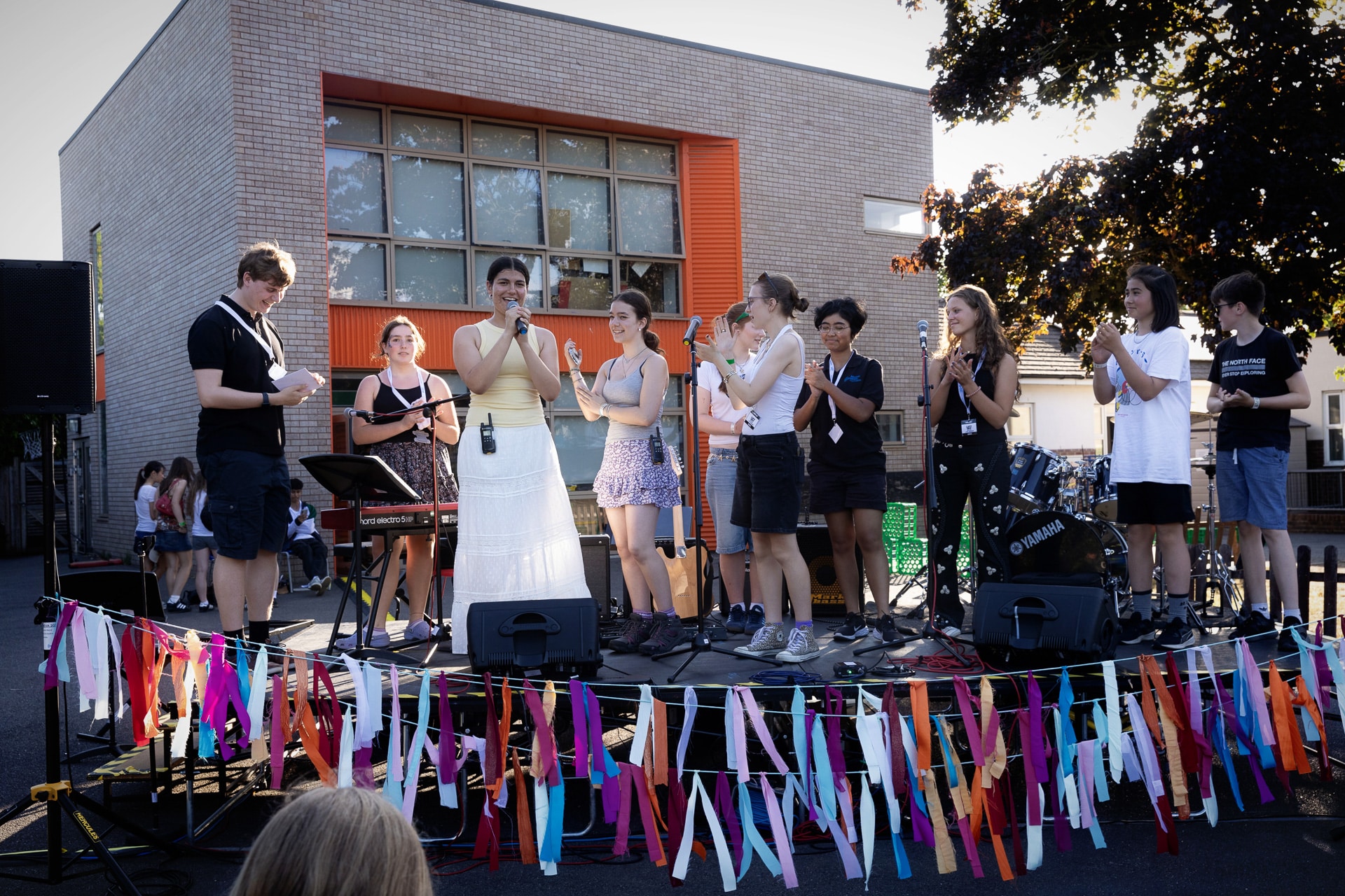 A group of young people stand on an outdoor stage, clapping and smiling. Colourful ribbons decorate the stage, and musical instruments like a keyboard and drums are visible. A brick building is in the background.