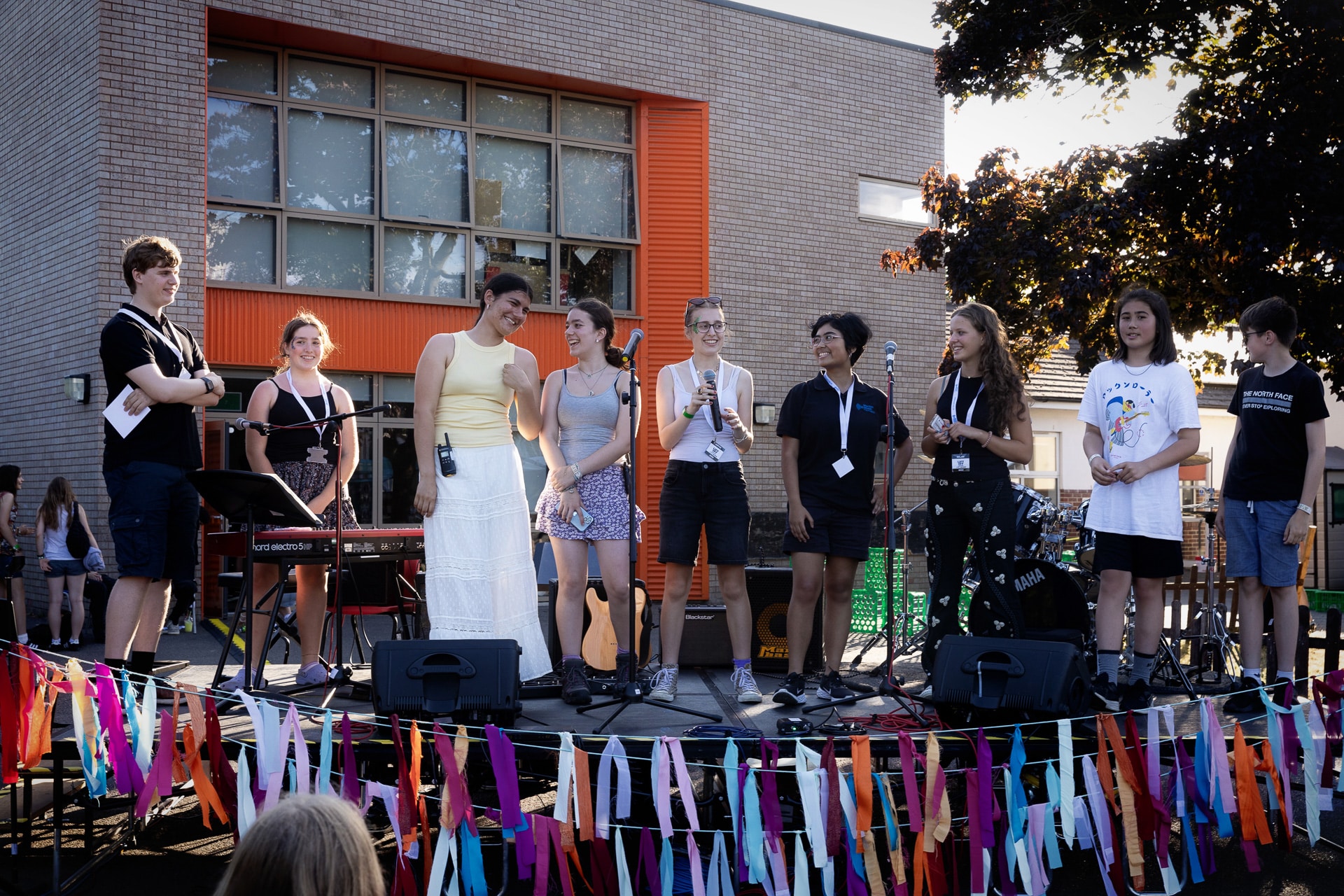A group of young people stand on an outdoor stage decorated with colourful streamers, holding microphones and instruments, in front of a modern brick building during a sunny event.
