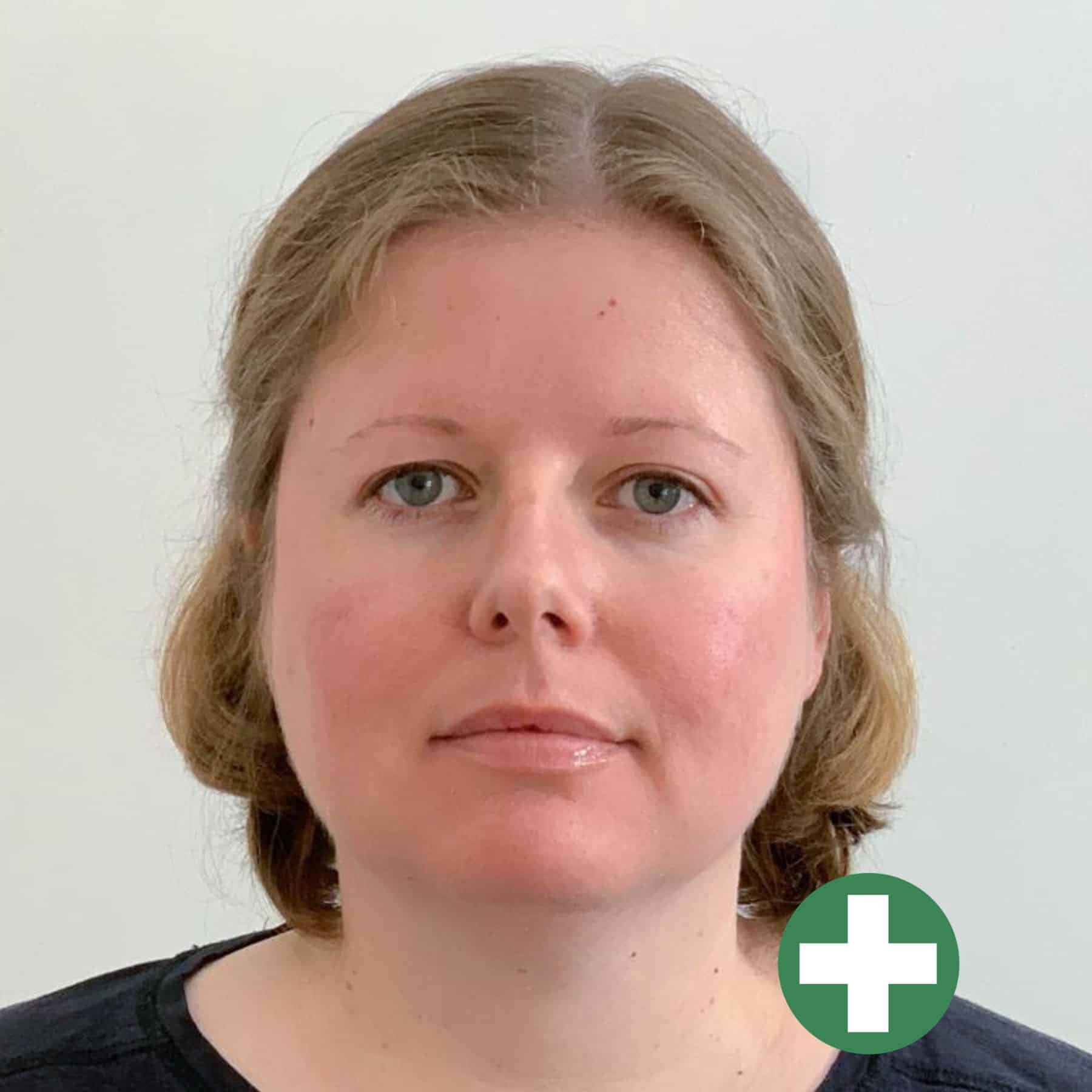 A woman with light skin and shoulder-length blonde hair is facing the camera with a neutral expression, wearing a dark top. A green circle with a white plus sign is at the bottom right corner of the image.