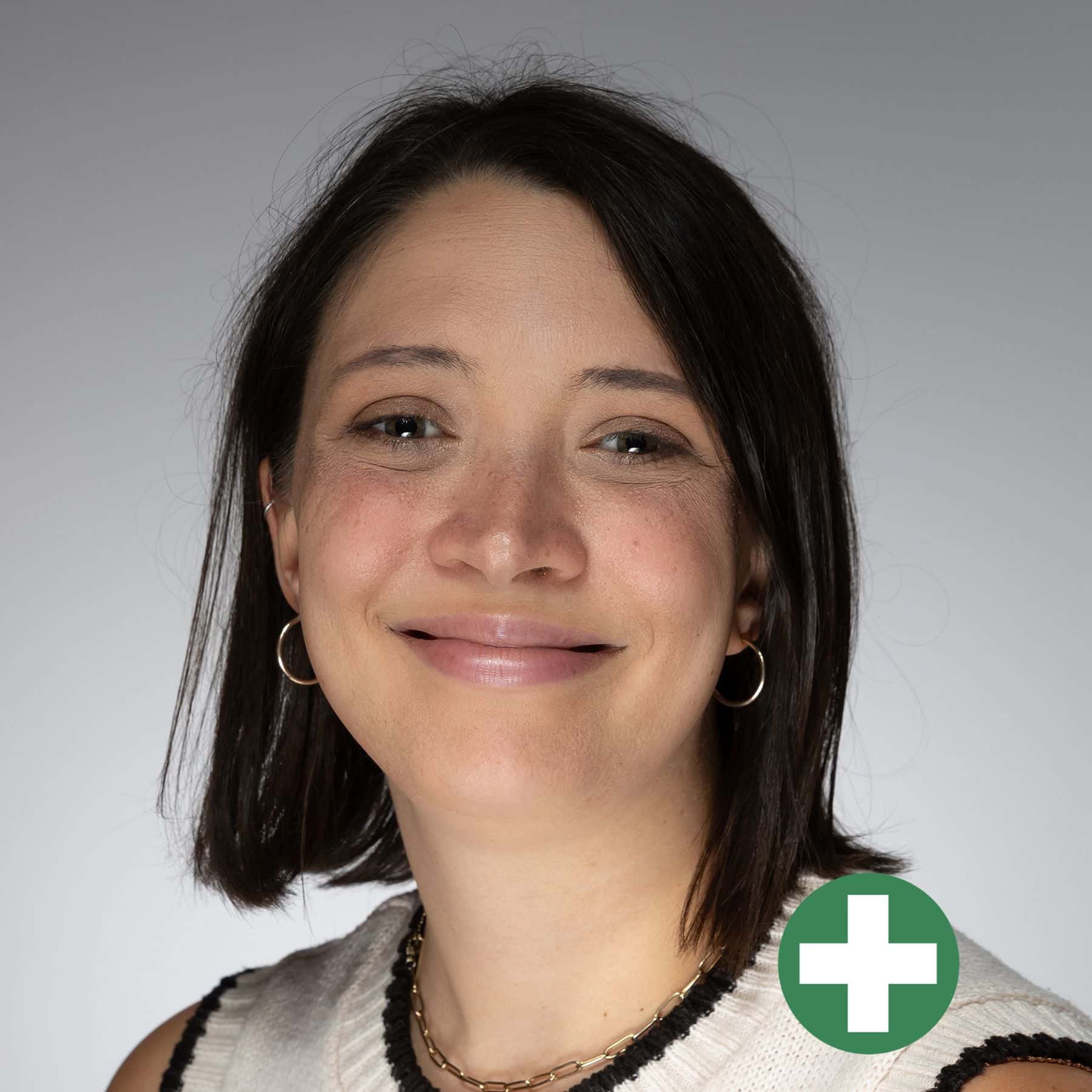 A woman with shoulder-length dark hair and hoop earrings smiling at the camera, wearing a sleeveless white top. There is a green circle with a white plus sign at the bottom right corner of the image.
