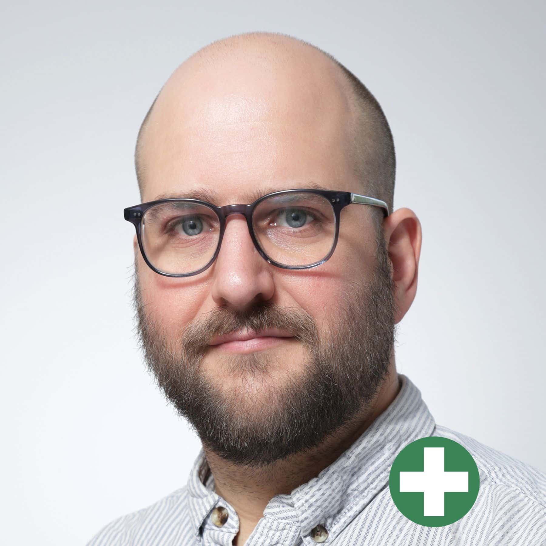 A bearded man with glasses and a bald head is wearing a light striped shirt, looking at the camera. In the bottom right corner, there is a green circle with a white plus sign inside.