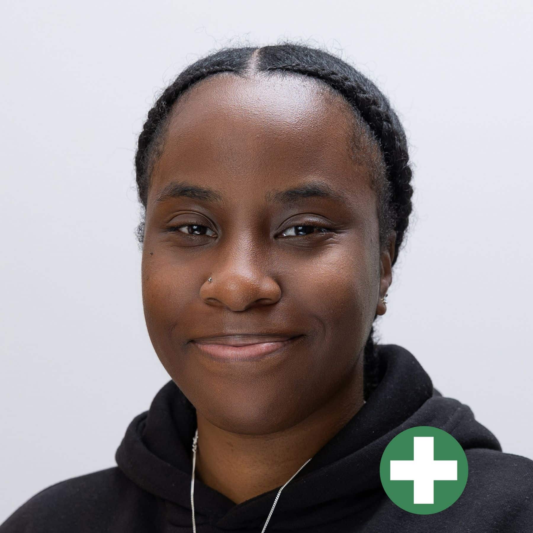 A person with plaited hair, wearing a black hoodie, smiles softly at the camera. There is a green circle with a white plus sign at the bottom right corner of the image.