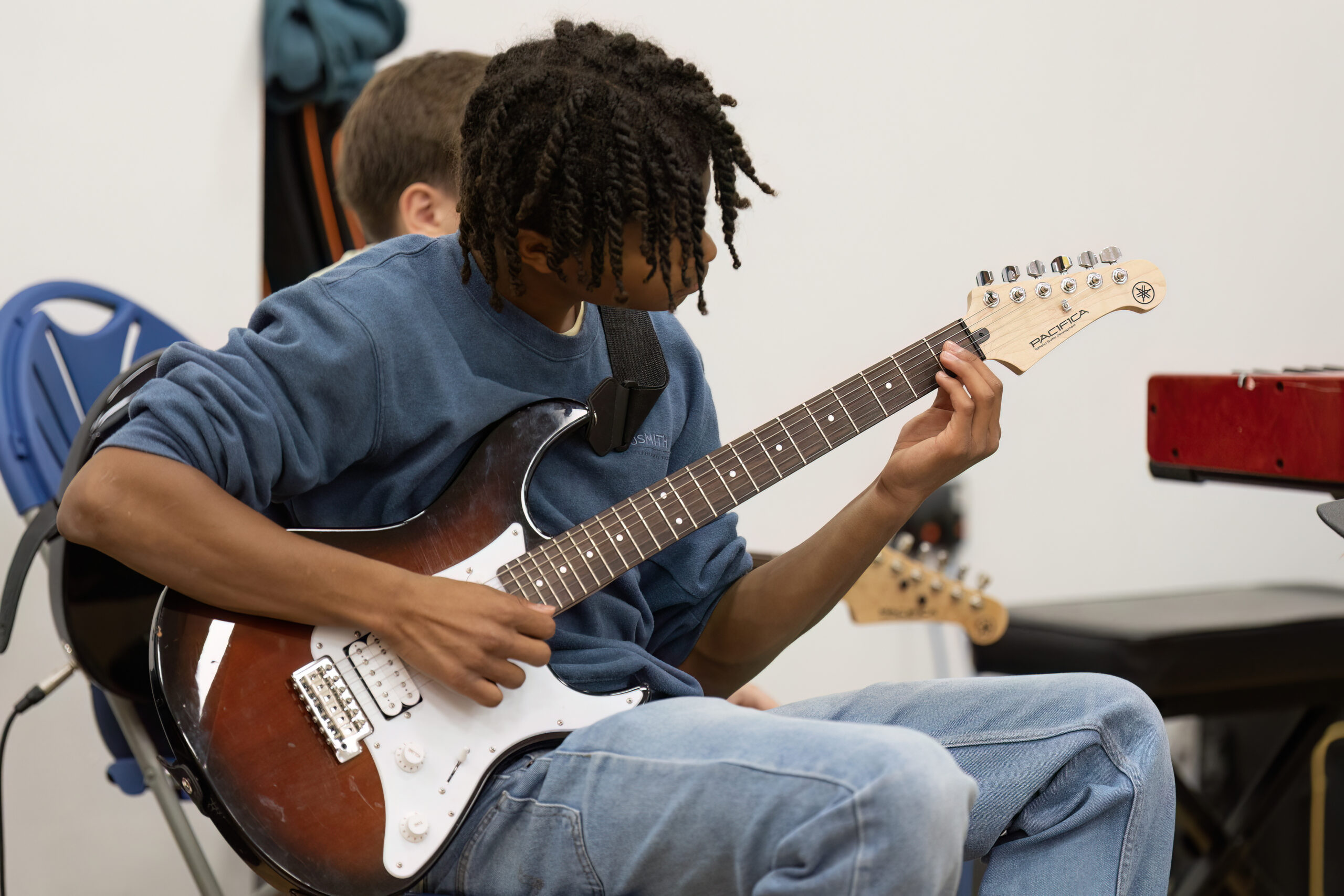 A young person with plaited hair plays an electric guitar, seated and focused, wearing a blue jumper and jeans. Another person sits partially behind them, with musical equipment visible in the background.