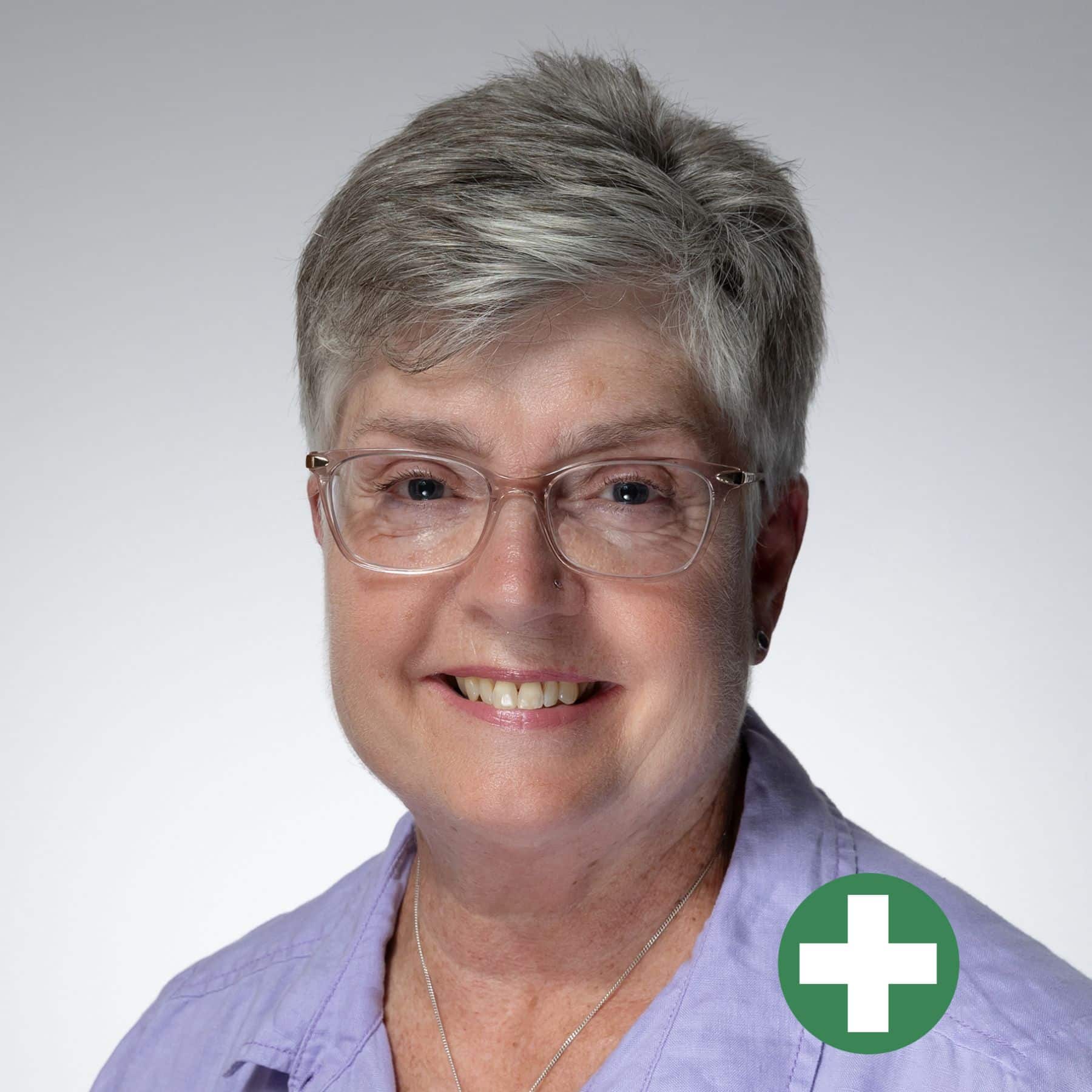 Smiling older woman with short grey hair and glasses, wearing a light purple shirt. A green circle with a white plus sign is overlaid in the lower right corner of the image.