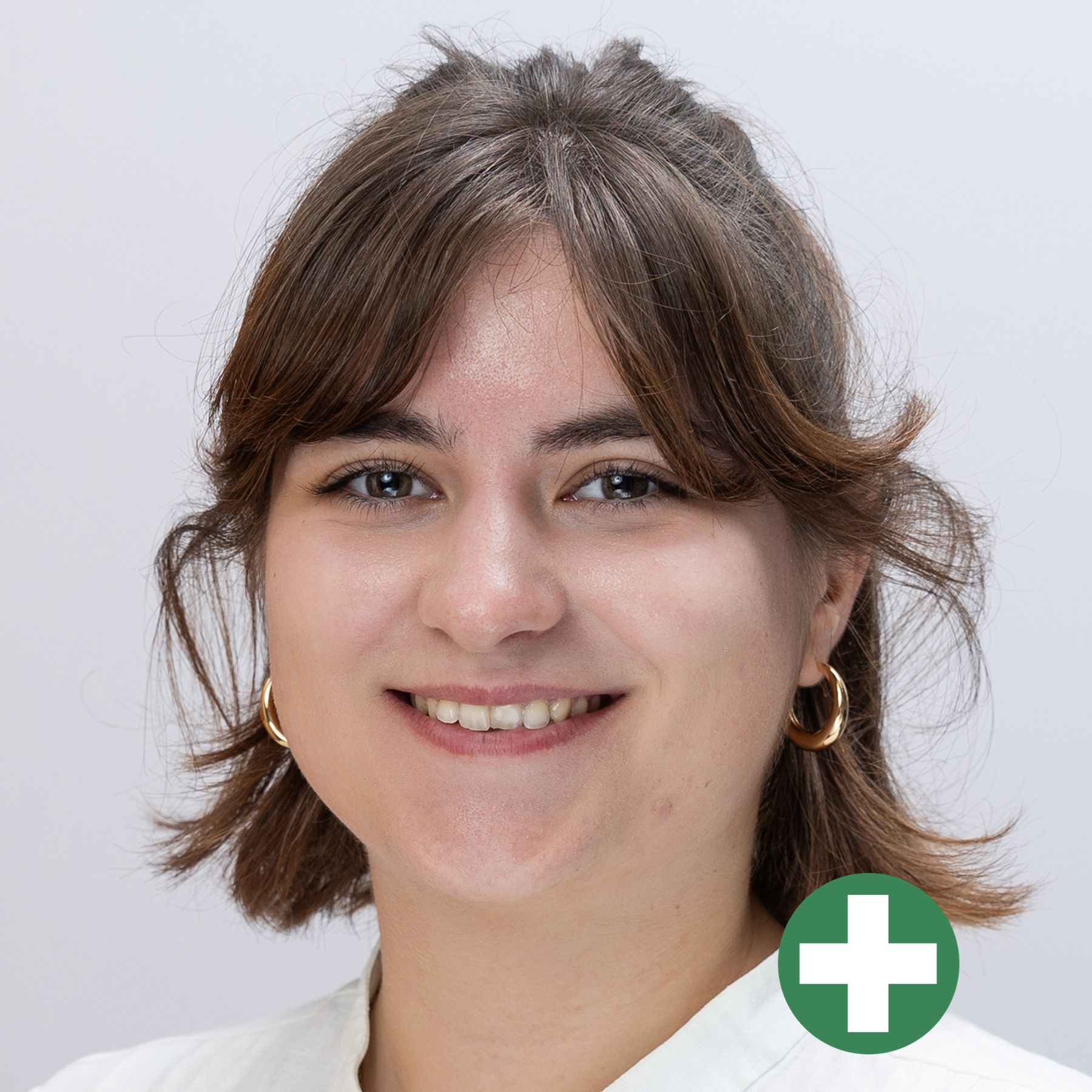 A young woman with short brown hair and gold hoop earrings smiles at the camera. She is wearing a light-coloured top. In the lower right corner, there is a green circle with a white plus symbol.