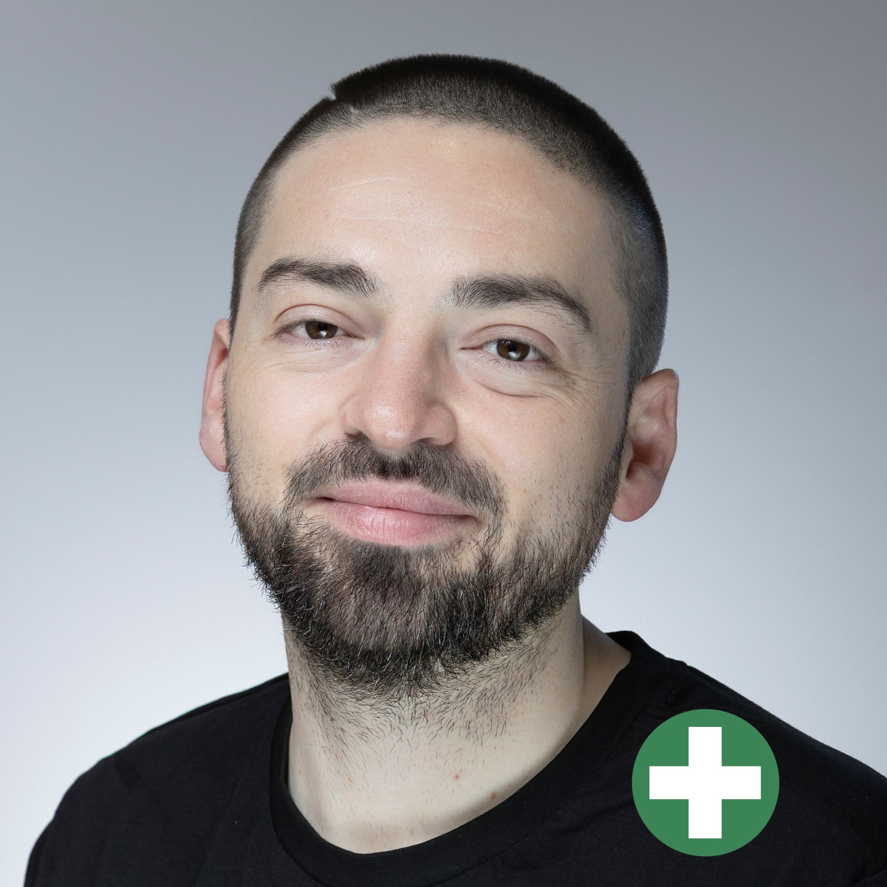 A man with short dark hair and a trimmed beard smiles at the camera. He is wearing a black shirt. There is a green circle with a white plus sign in the bottom right corner of the image.