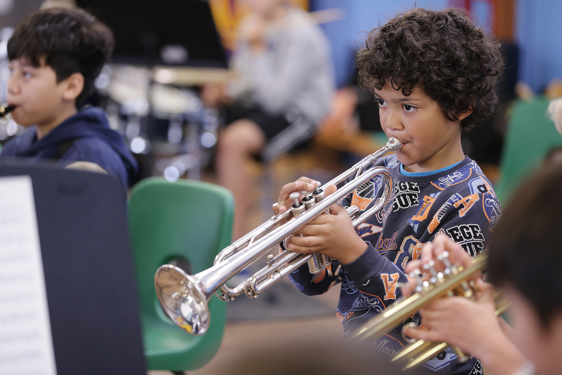 A young boy with curly hair plays the trumpet during instrumental lessons in a music classroom, surrounded by other children playing instruments. He is focused, wearing a long-sleeve shirt with varsity-style lettering.