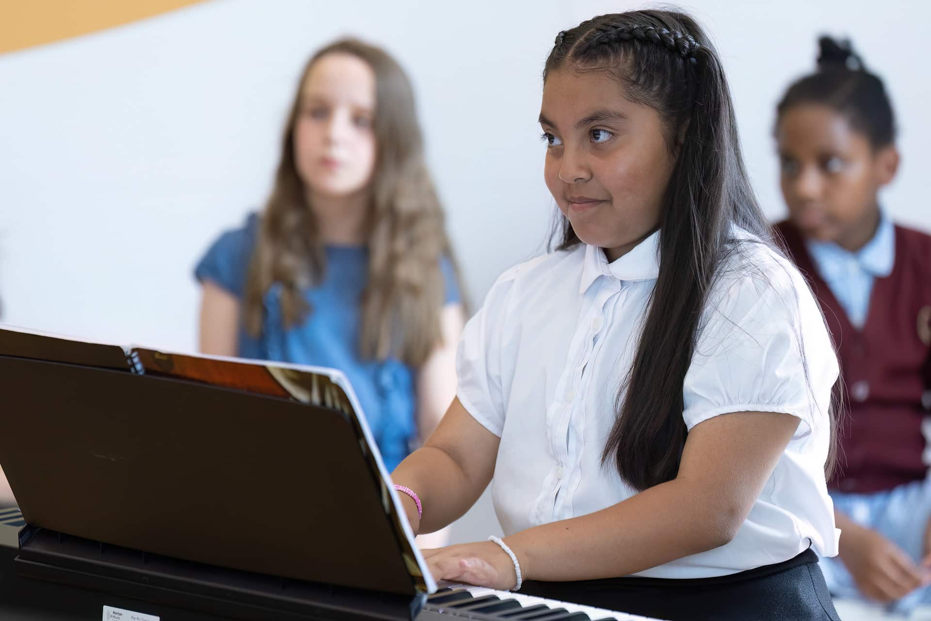 A girl with long dark hair plays a keyboard during her instrumental lessons, reading sheet music, whilst two other girls sit in the background, slightly out of focus.