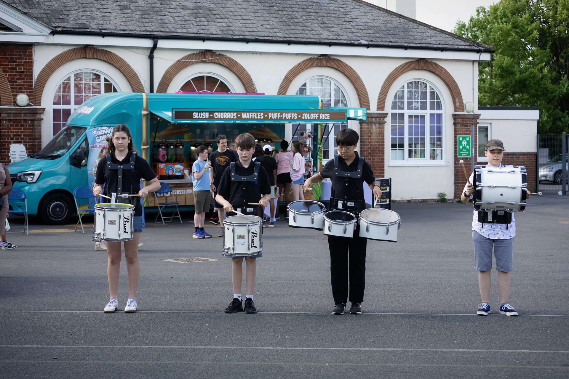 Four young people stand in a row outdoors, each playing a marching snare drum—perhaps showing off skills from their instrumental lessons. Behind them is a turquoise food van and a building with arched windows, with several people gathered nearby.
