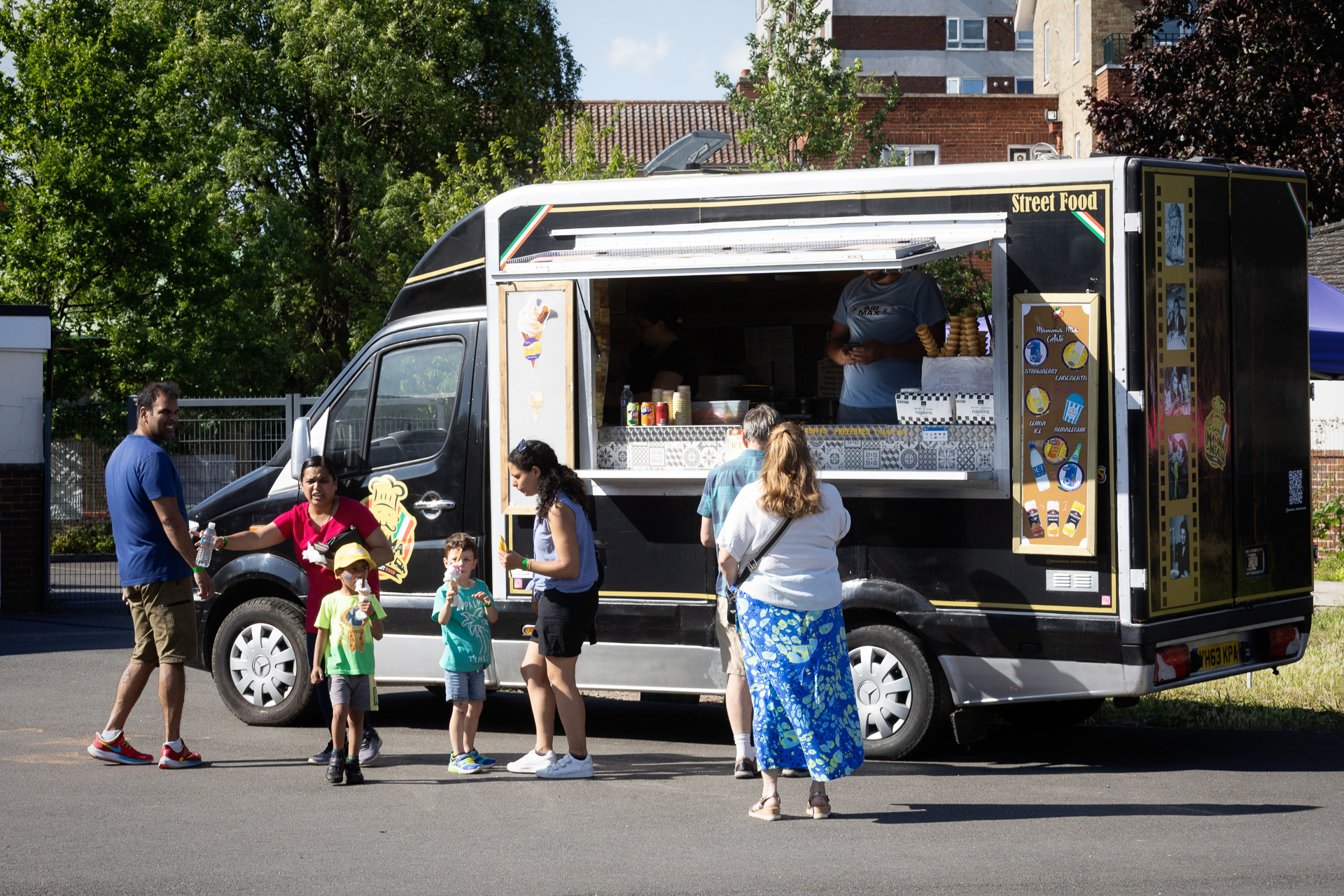 A group of people, including adults and children, stand in front of a black food van with a serving window, waiting for or receiving food on a sunny day in an outdoor urban setting after their instrumental lessons.