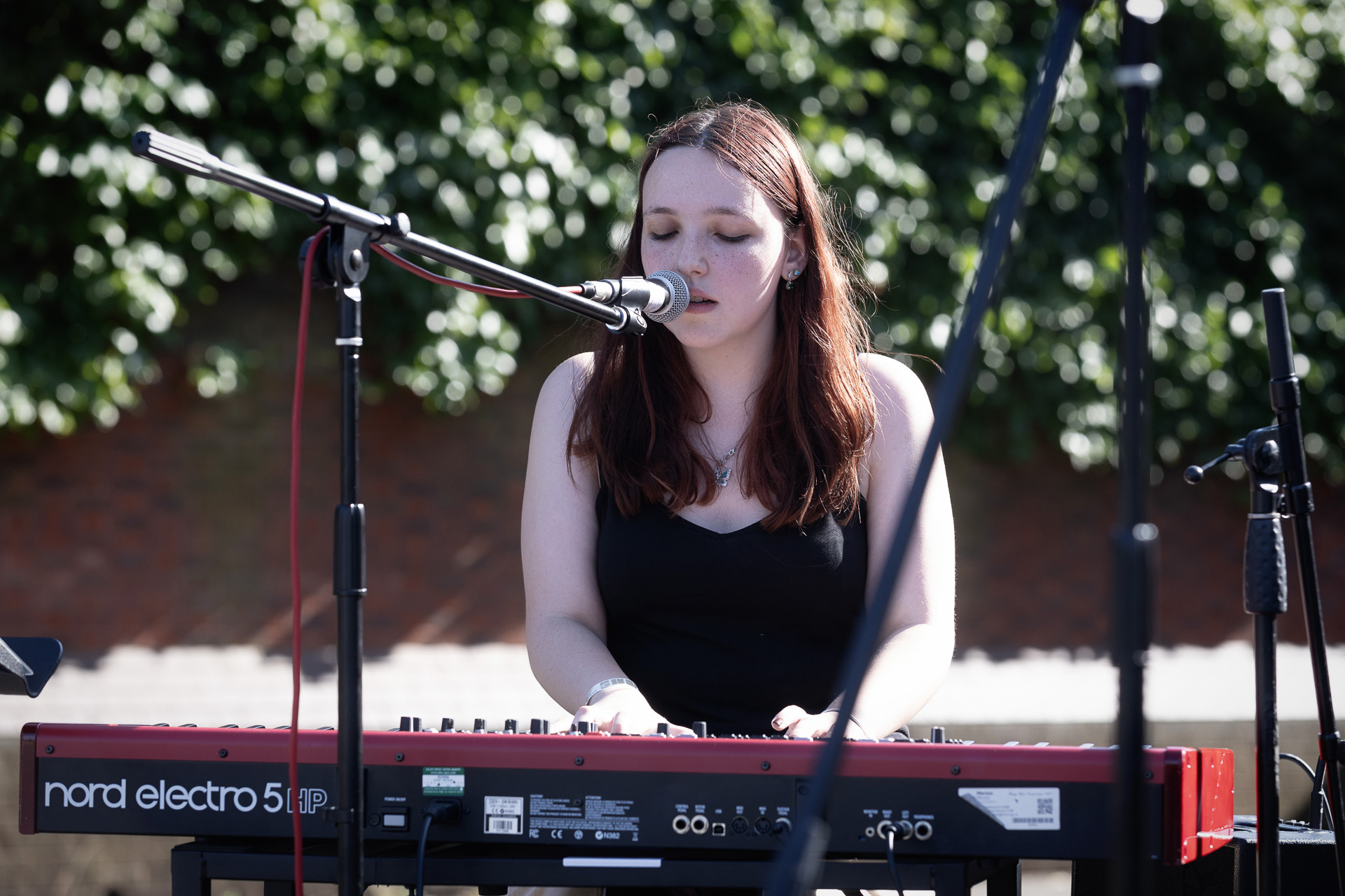 A young woman with long brown hair plays a red Nord keyboard during instrumental lessons and sings into a microphone outdoors, with greenery and a brick wall in the background. She wears a black vest top and appears focused on her performance.