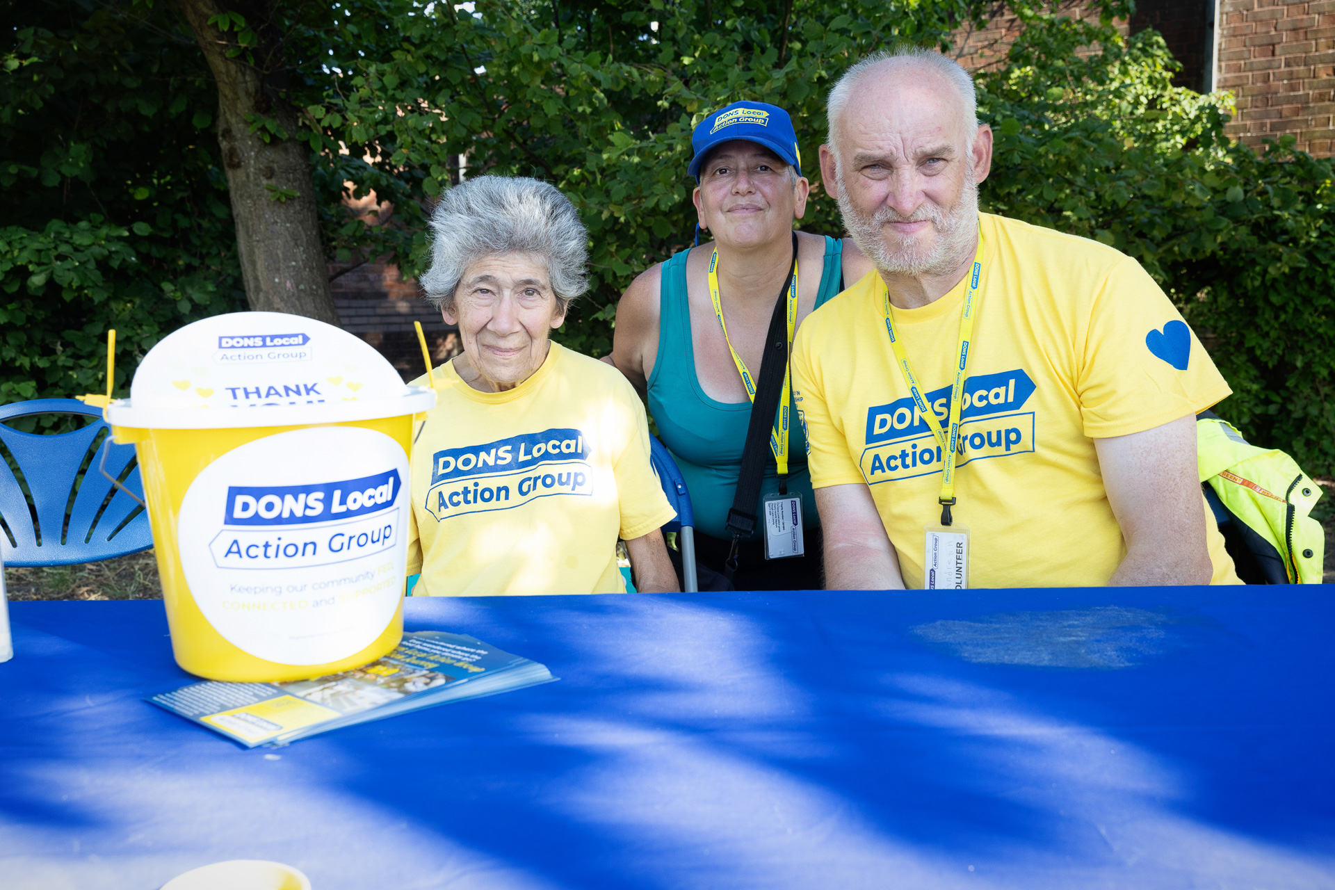 Three people from the DONS Local Action Group, in yellow logo shirts, sit at a table with a donation bucket and leaflets on a blue cloth at the Lift Off Festival. Trees and a brick building can be seen in the background.