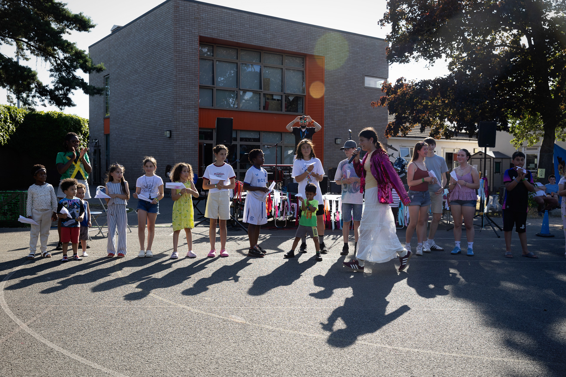 A group of children and adults stand in a queue outdoors, some holding papers, as a person in a bright pink outfit walks by. The festive scene suggests they're at the Lift Off Festival, with sunlight streaming past a building in the background.