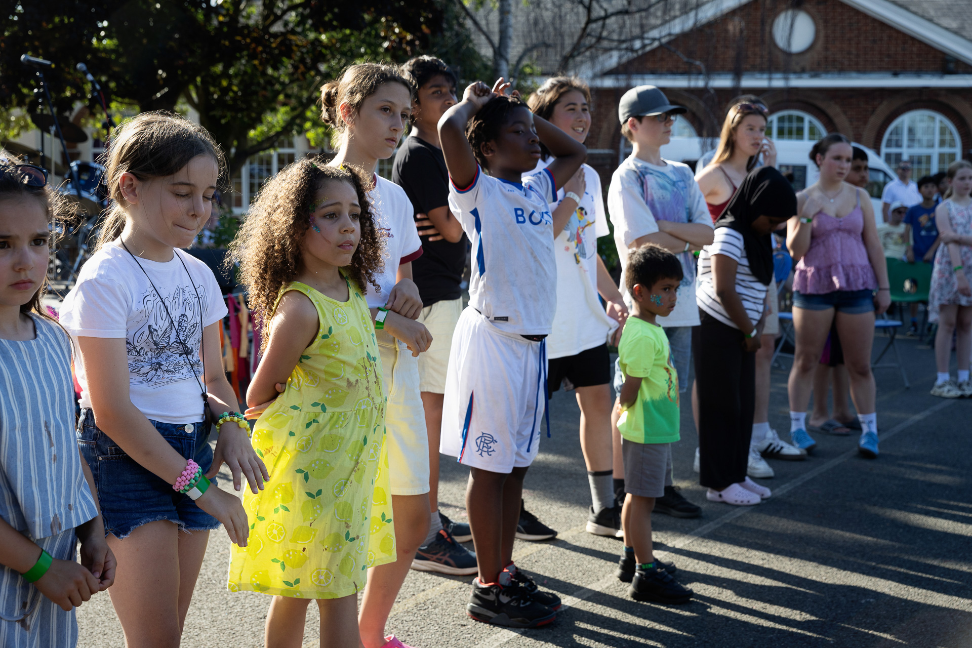 A group of children stand outdoors in sunlight, some with painted faces, watching something off-camera. They are casually dressed in summer clothes, possibly gathered for Instrumental Lessons, with a brick building and others visible in the background.