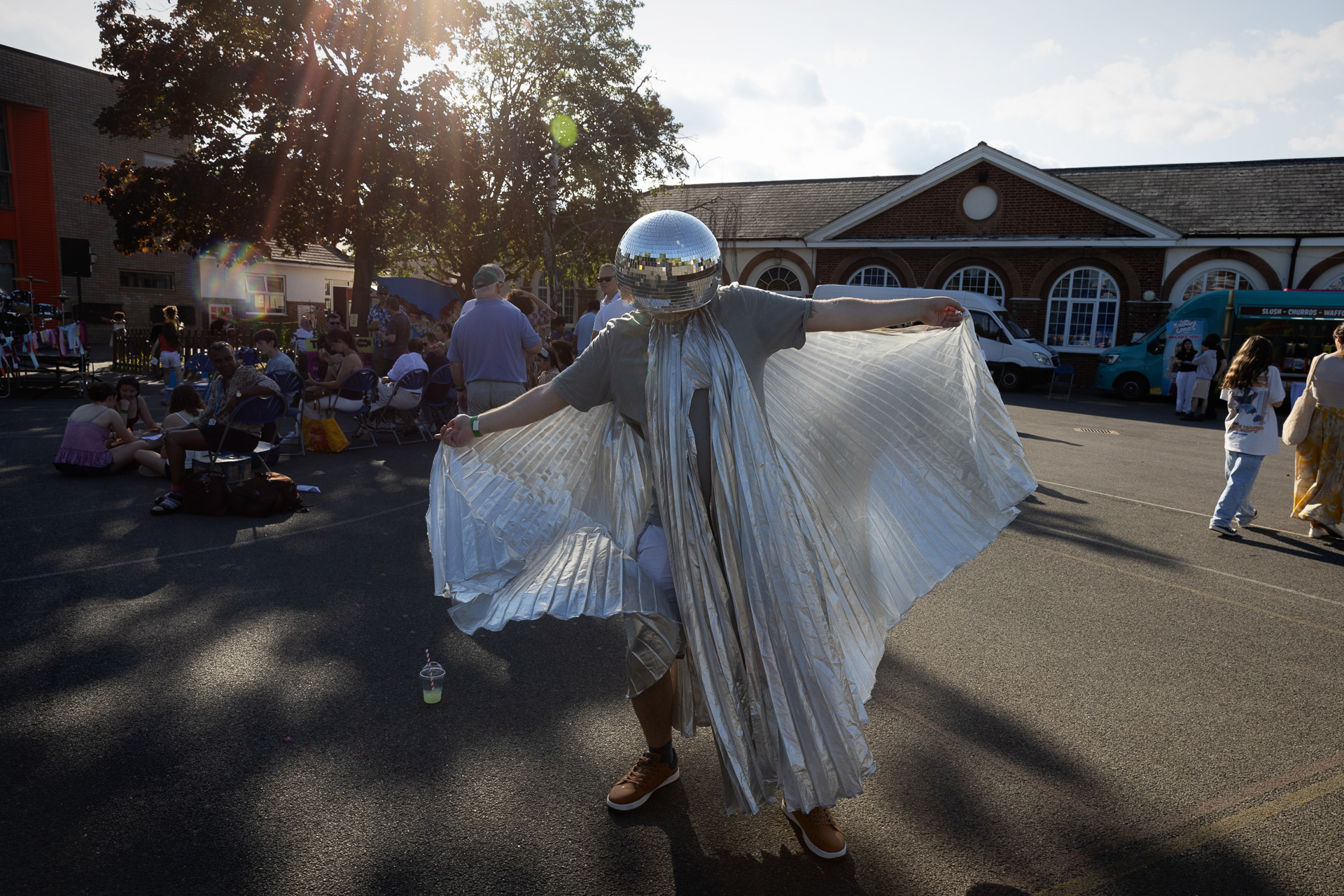 A person wearing a reflective disco ball helmet and a flowing silver cape poses with outstretched arms at the Lift Off Festival, with people and tents in the background under the afternoon sun.