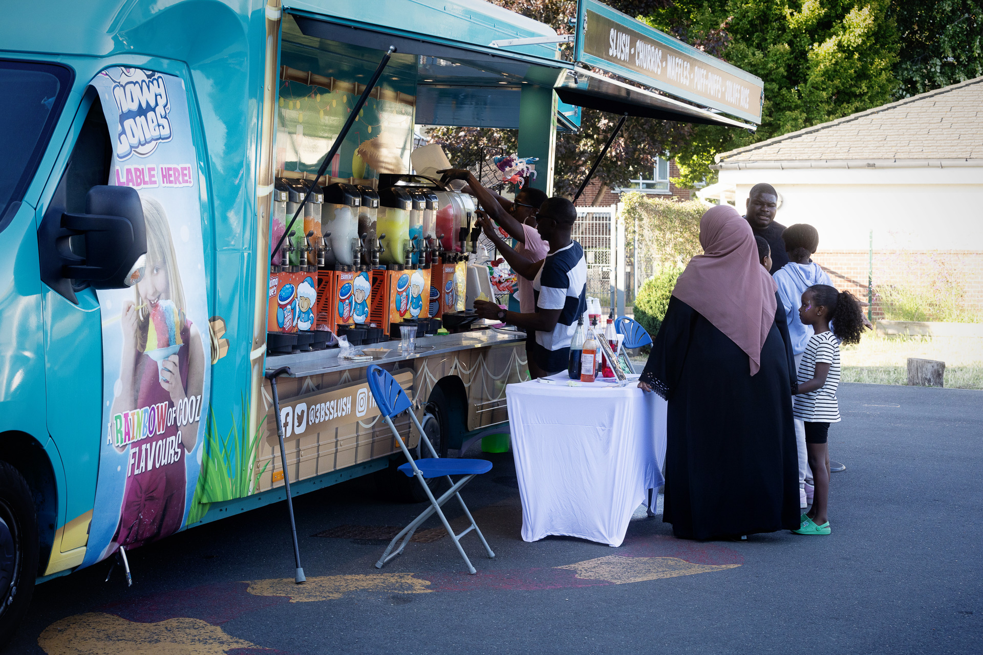 A group of people, including children, stand at a colourful food van at the Lift Off Festival, selling drinks with large dispensers. One person dispenses a drink while others wait. The van is parked outdoors on a sunny day.