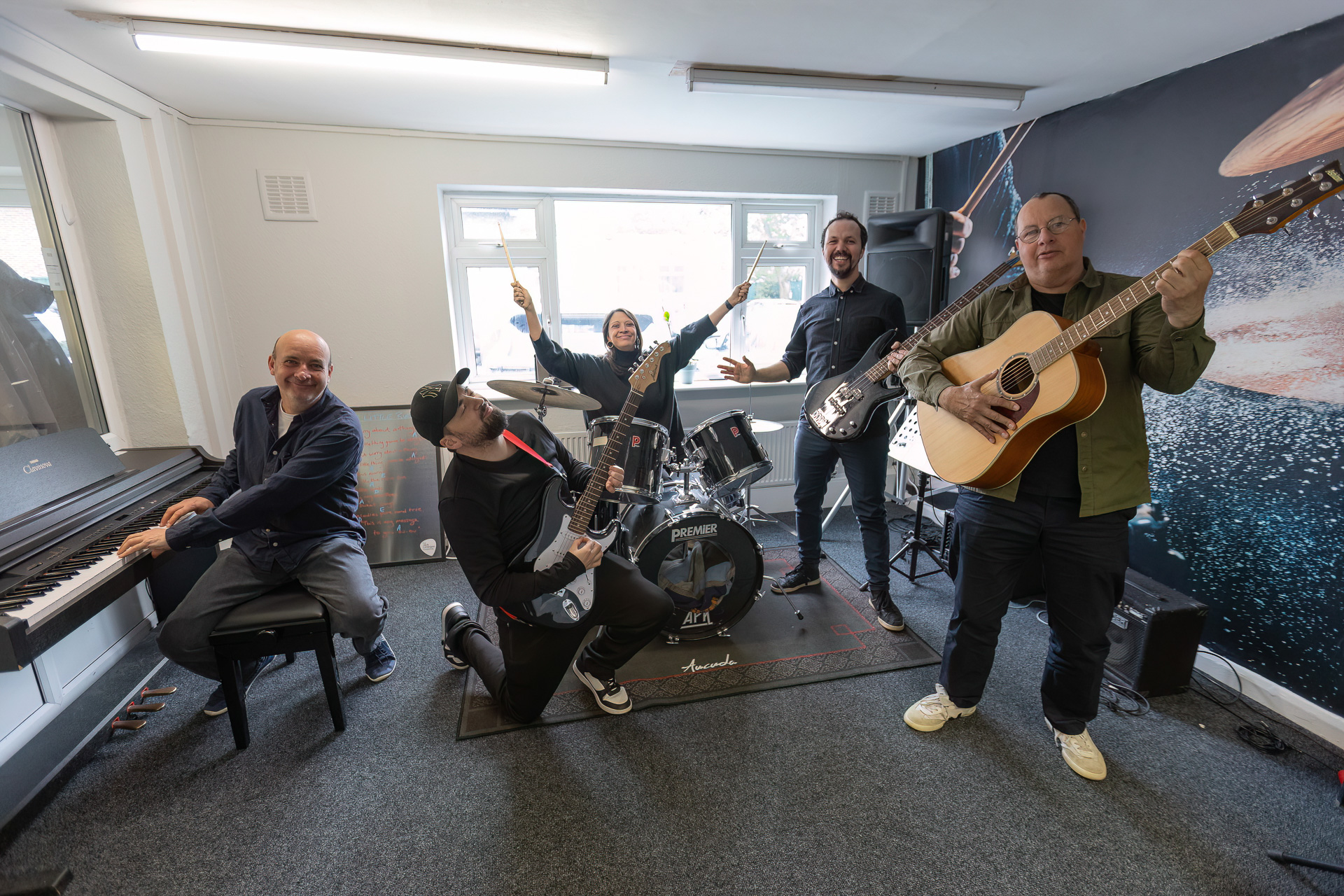 Five people, MMF and JLF stafff, enthusiastically pose as a band in a practice room with a drum kit, keyboard, electric guitars, and an acoustic guitar; all are smiling and appear to be having fun.