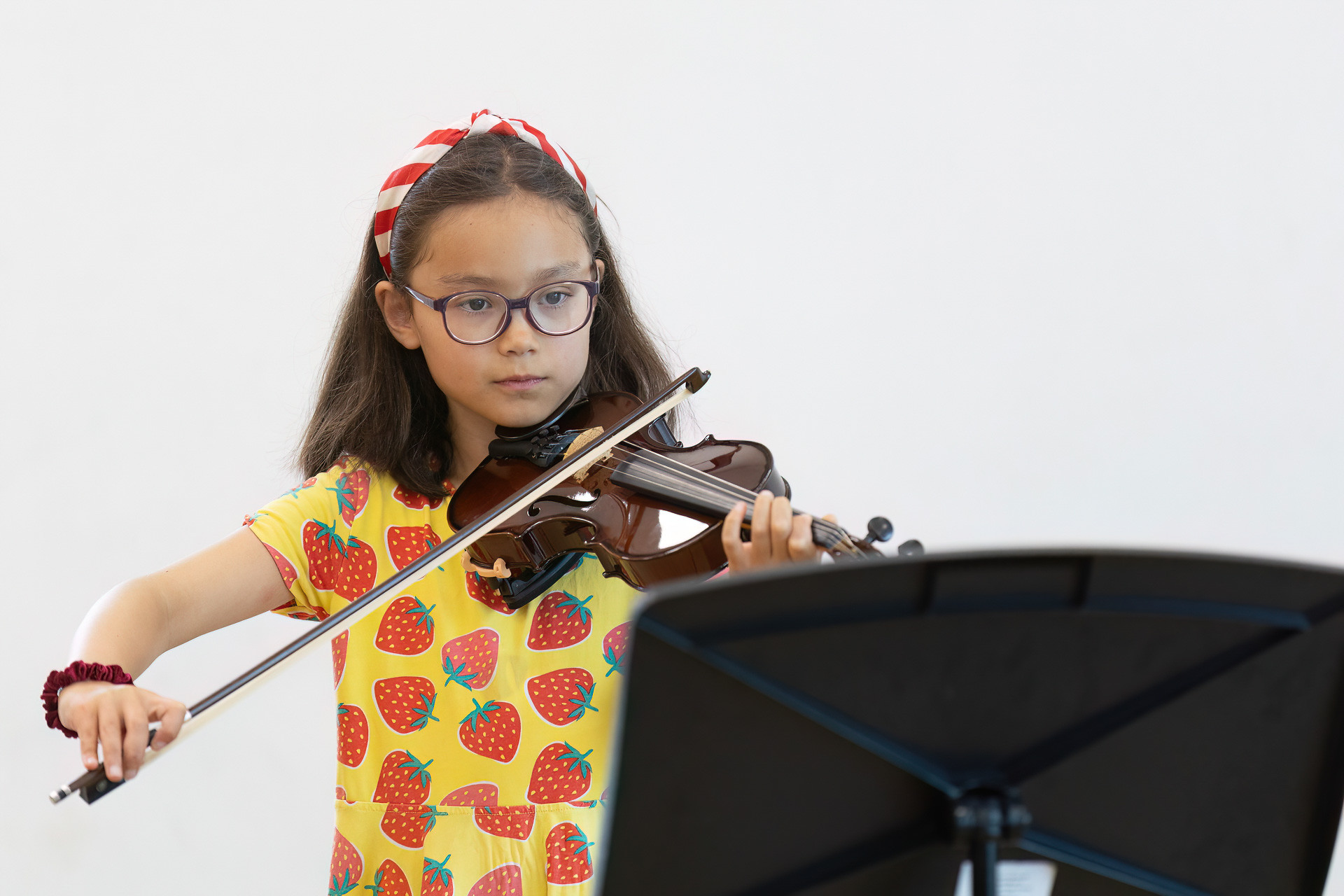 54751069507_c358a97b43_k A young girl wearing glasses and a strawberry-patterned dress plays the violin during her instrumental lessons, looking at sheet music on a stand in front of her. She has a red and white striped hairband in her hair.