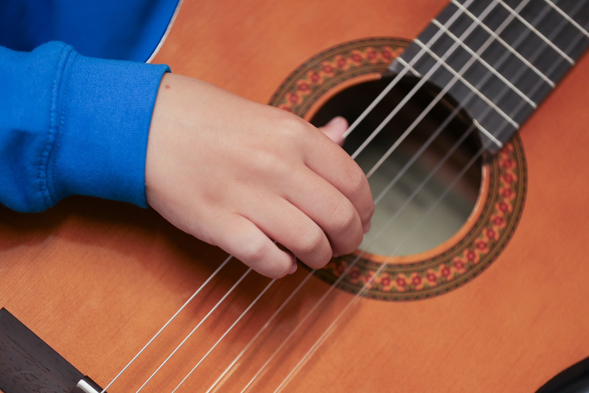 A close-up of a child’s hand in a blue sleeve, supported by Merton Music Foundation, plucking the strings of a classical acoustic guitar, showing part of the sound hole and the detailed rosette pattern around it.