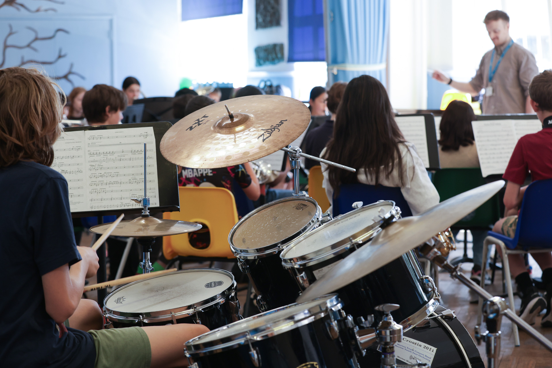 A student plays a drum kit in a classroom with other Merton Music Foundation students on instruments. Music sheets are on stands, and a teacher conducts at the front, creating a bright and focused atmosphere.