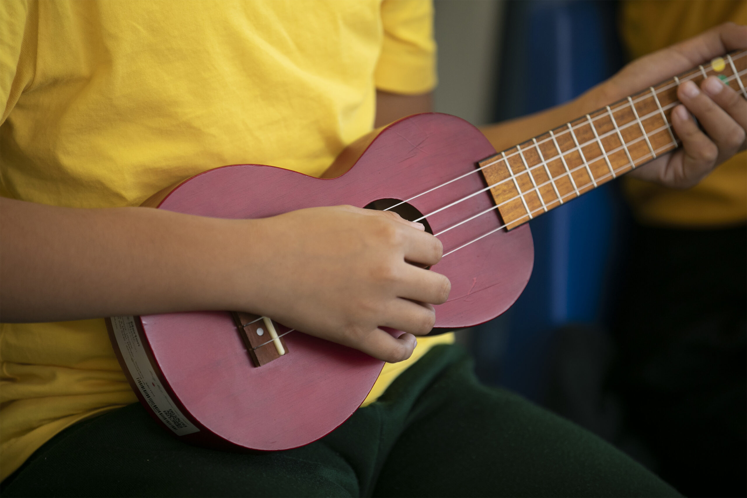 A person wearing a yellow shirt and dark green trousers is playing a small, pink ukulele, strumming with one hand and holding the neck with the other—perfect for illustrating beginner Instrumental Lessons with focus on technique.
