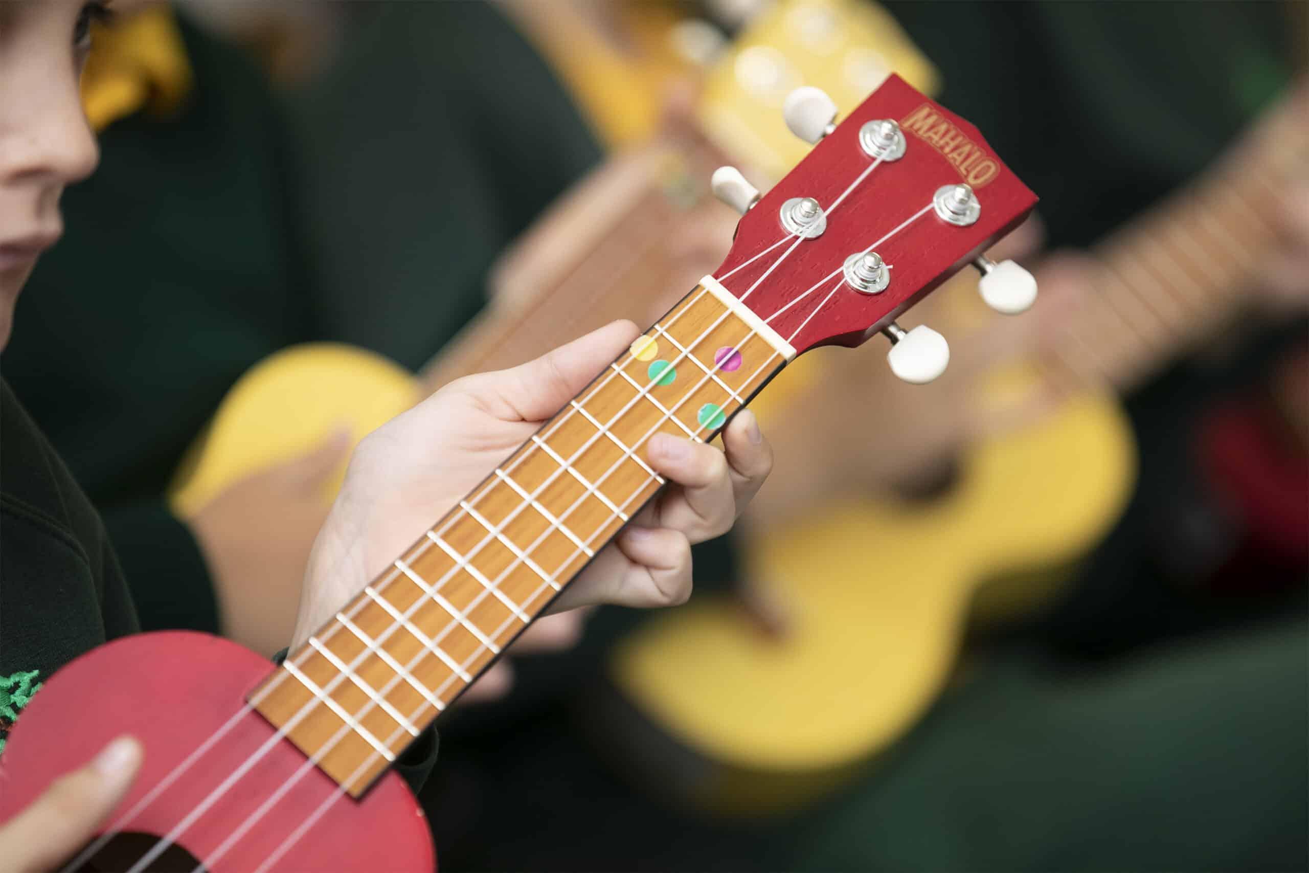 Close-up of a person playing a red ukulele during instrumental lessons, focusing on their hand pressing the strings on the fretboard. Several people with yellow ukuleles are visible in the blurred background.