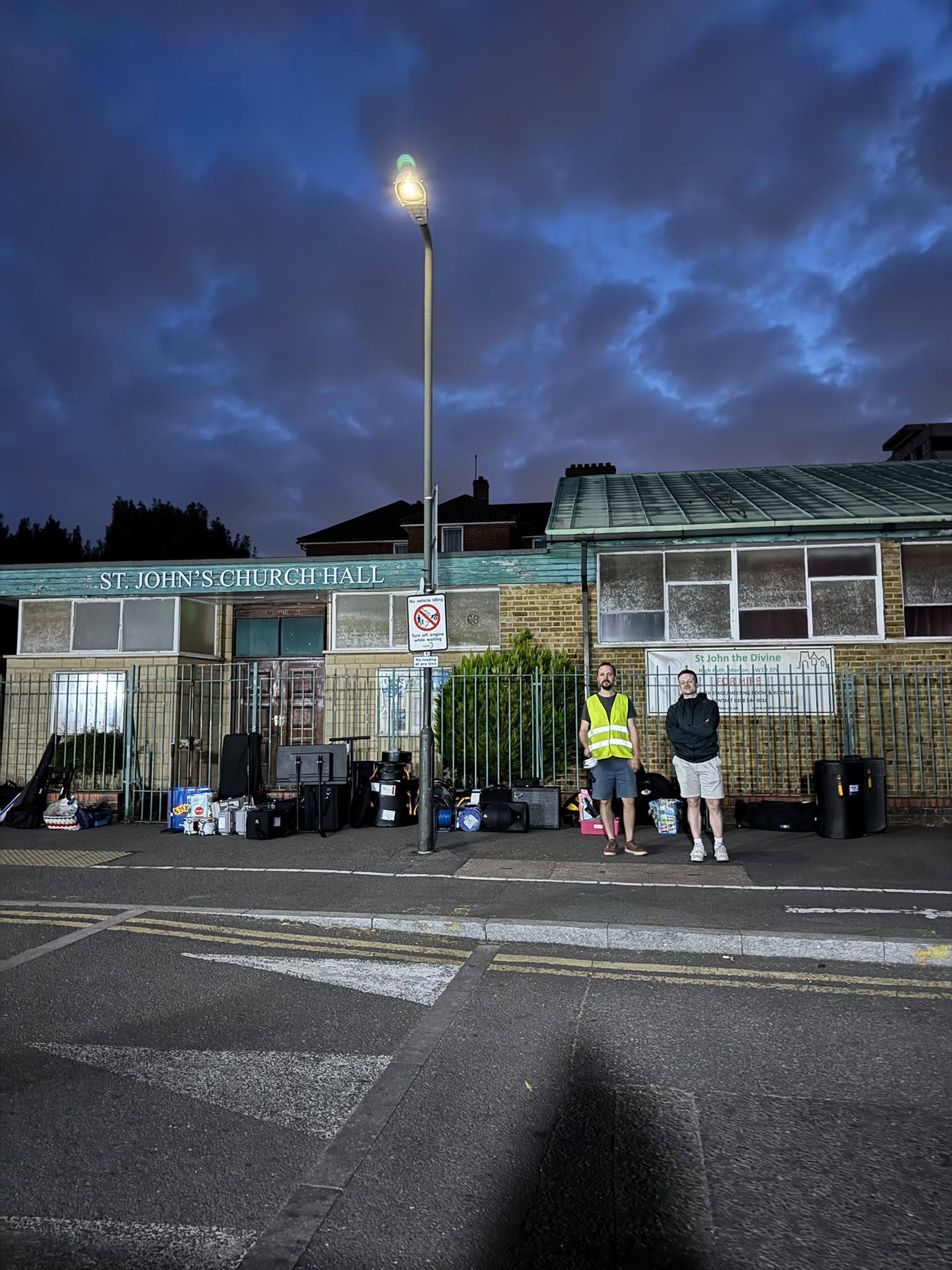 Two new TEMPO Ambassadors stand on a pavement at night near St. Johns Church Hall, surrounded by bags and supplies, under a streetlamp with a metal fence and cloudy blue sky in the background.