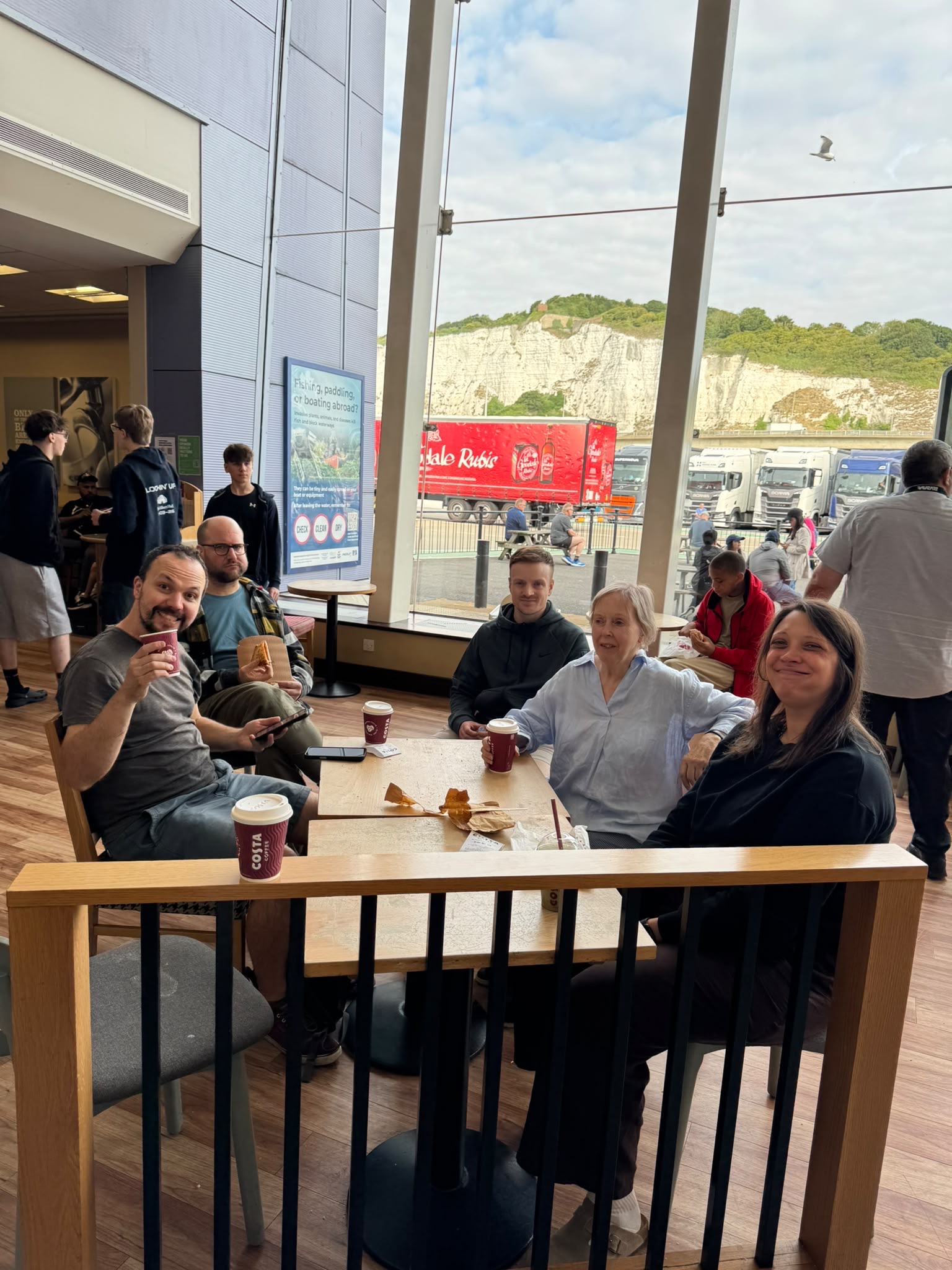 Five people sit around a table in a café with coffee and snacks. Large windows behind them show cliffs, vehicles, and people outside. The mood appears casual and relaxed, as the group of New TEMPO Ambassadors enjoys their conversation and camaraderie.