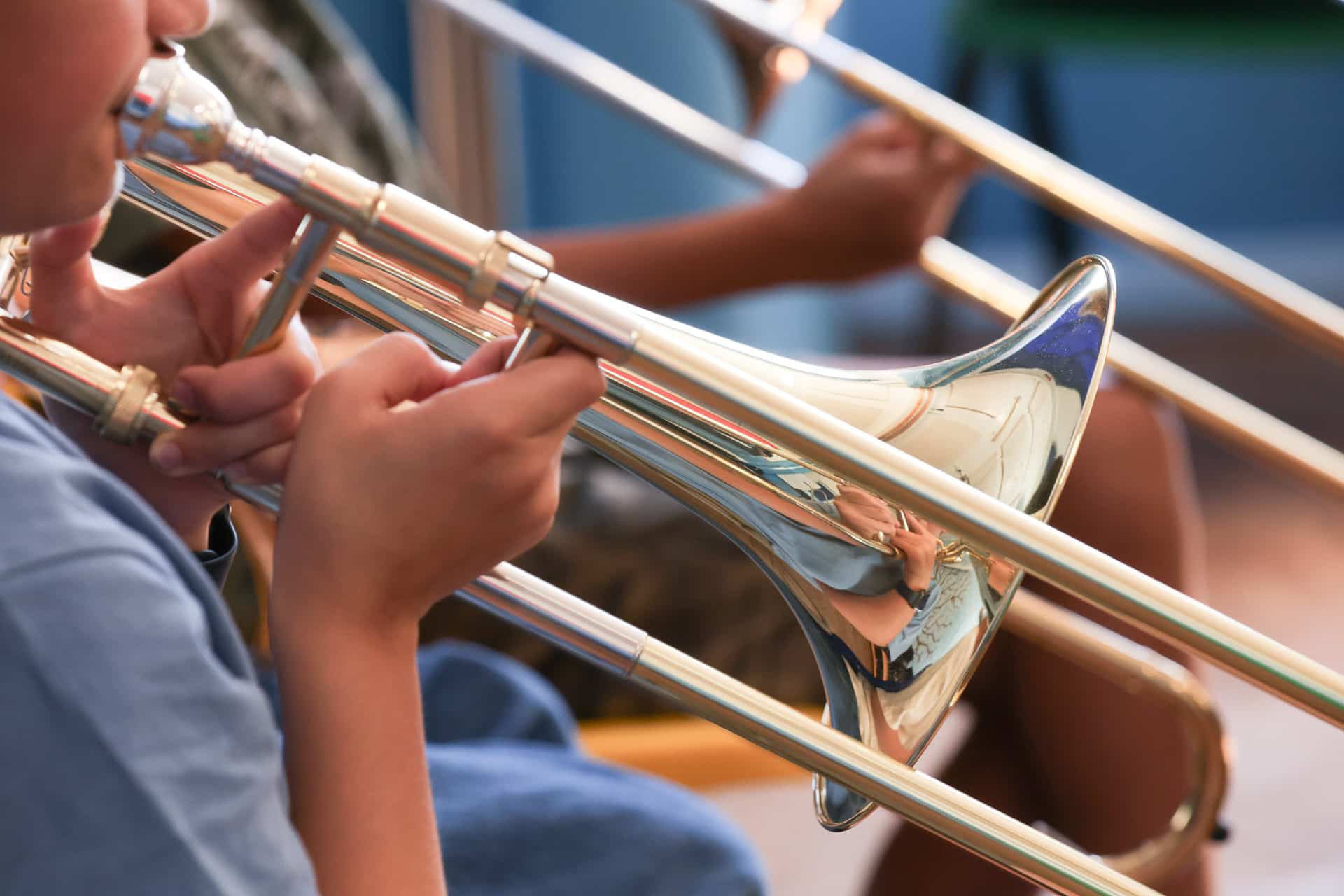 A close-up of two people playing shiny brass trombones, focusing on their hands and the instruments, with a blue background blurred in the distance—capturing a vibrant moment from a Merton Music Foundation event.