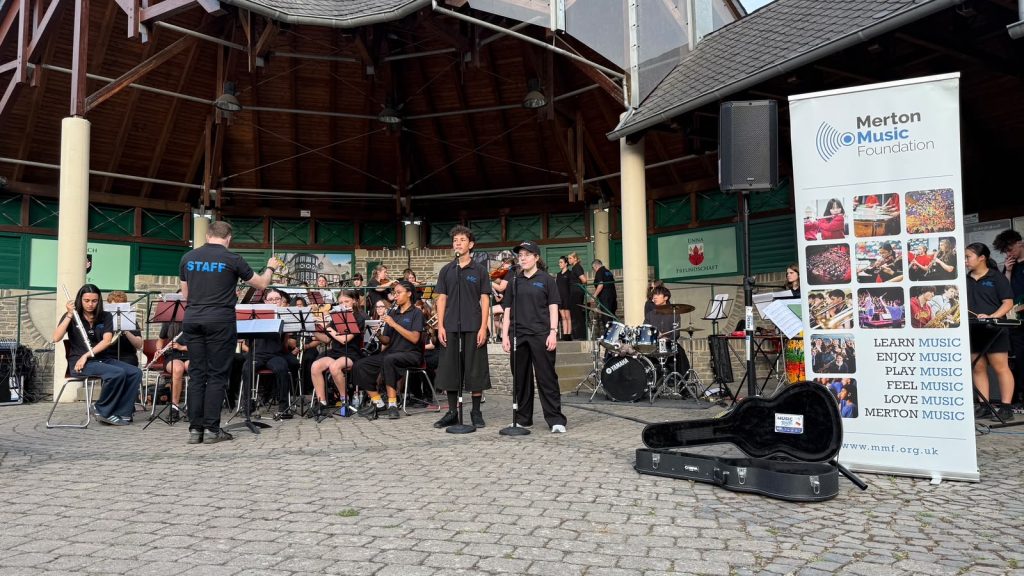 A band of young musicians performsoutdoors under a pavilion, with singers in front, a conductor, instruments, and a Merton Music Foundation banner on the right. The event appears lively and well-attended.