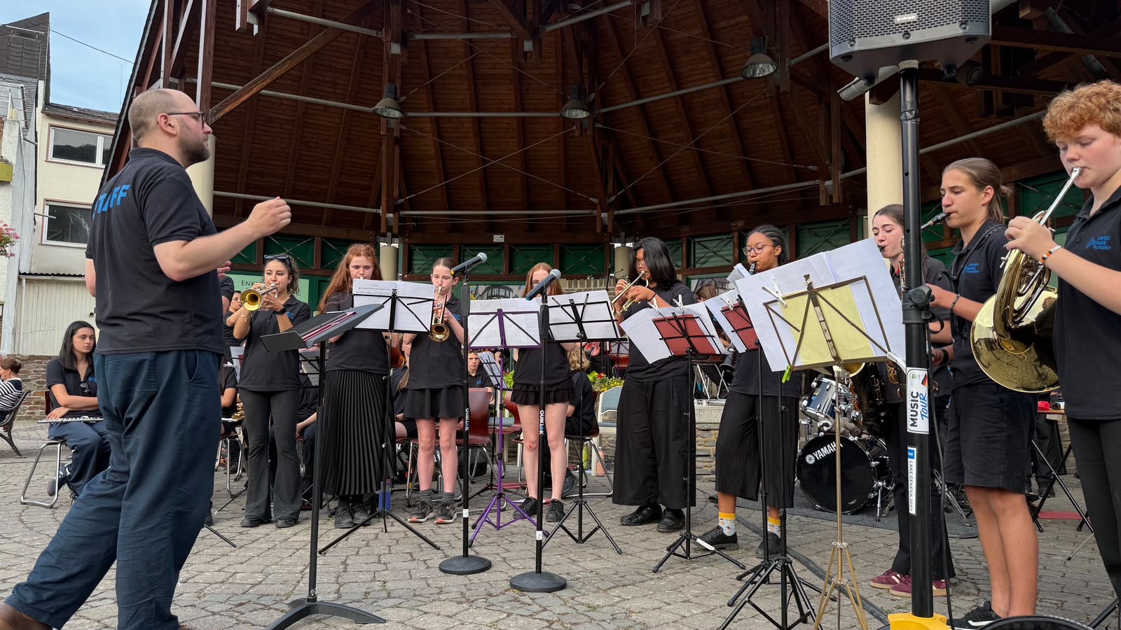 A conductor leads a group of young musicians, playing brass and woodwind instruments on an outdoor stage, with an audience seated nearby under a wooden pavilion.
