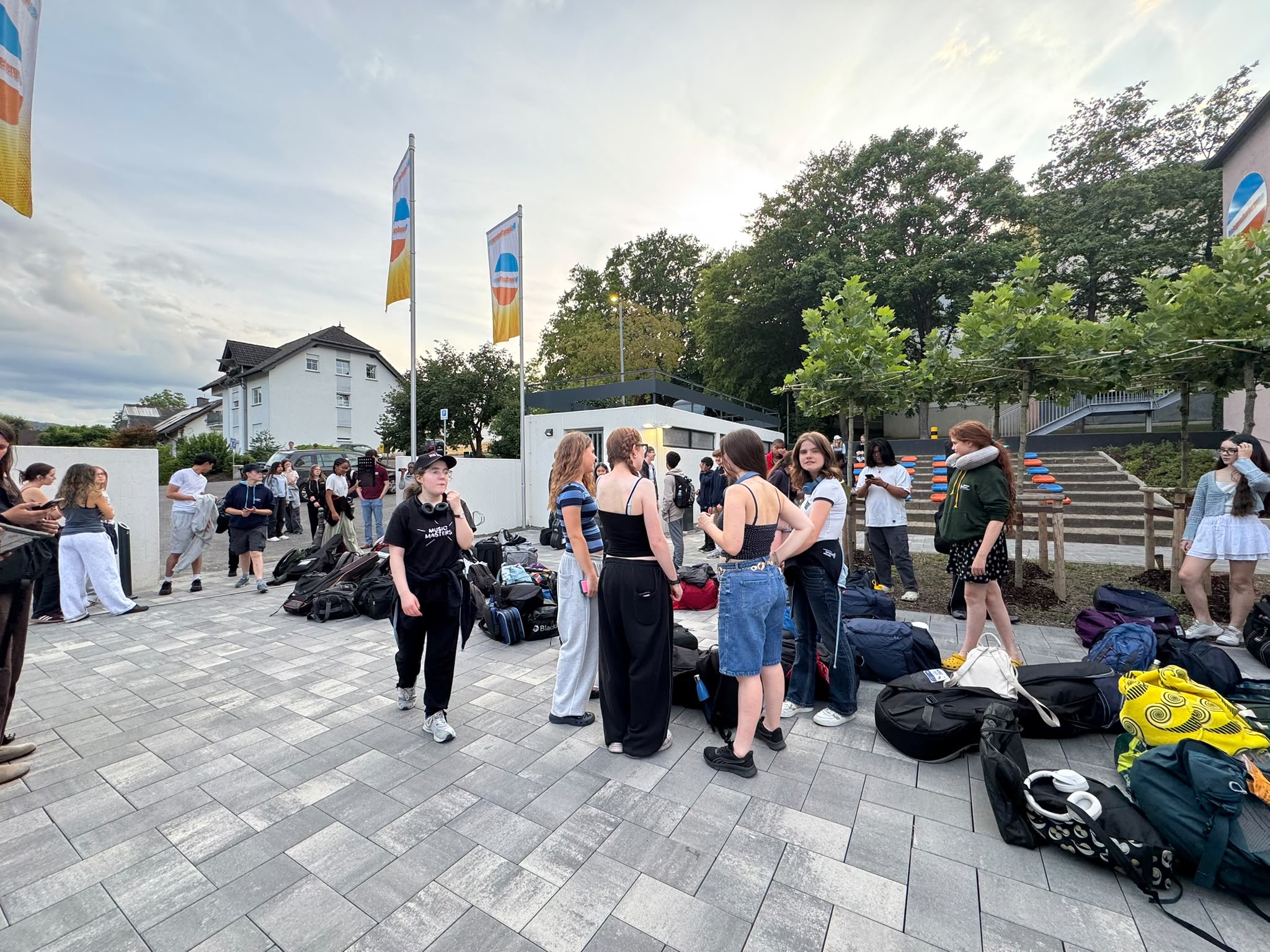 A group of young people, including the new TEMPO Ambassadors, stand and talk outside on a paved area, surrounded by bags and rucksacks, with flags and trees in the background.