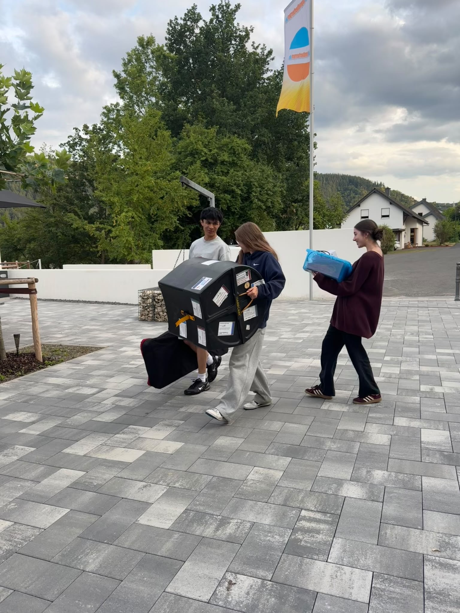 Three new TEMPO Ambassadors walk outdoors on a paved path. One person carries black boxes, another holds a blue item, and the third walks alongside them. Houses, trees, and a colourful flag are visible in the background.