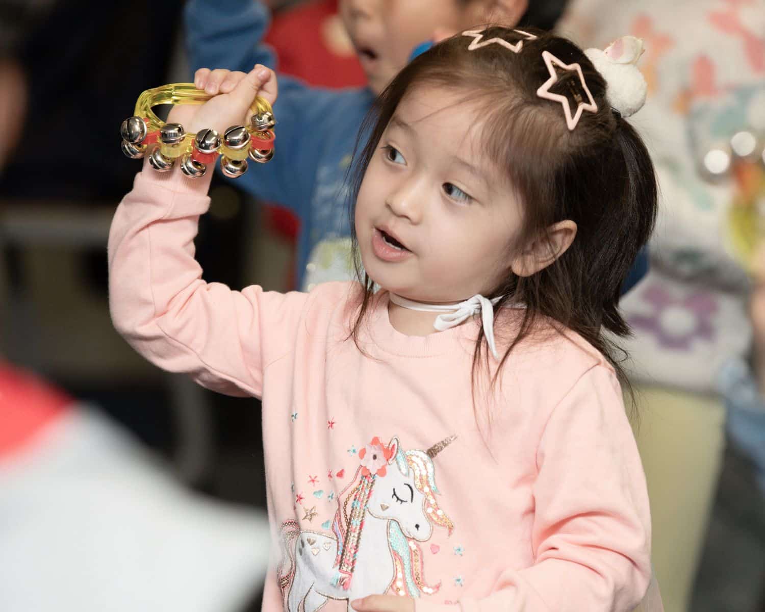 Young girl in a pink unicorn jumper, one of the new TEMPO Ambassadors, plays a yellow tambourine. She has star-shaped hair clips and looks to the side, surrounded by other children in a lively, indoor setting.