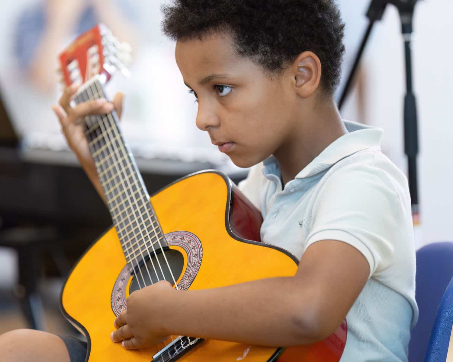 A young boy, one of the new TEMPO Ambassadors, sits and plays an acoustic guitar, looking focused. He wears a light-coloured polo shirt, and musical equipment is blurred in the background.