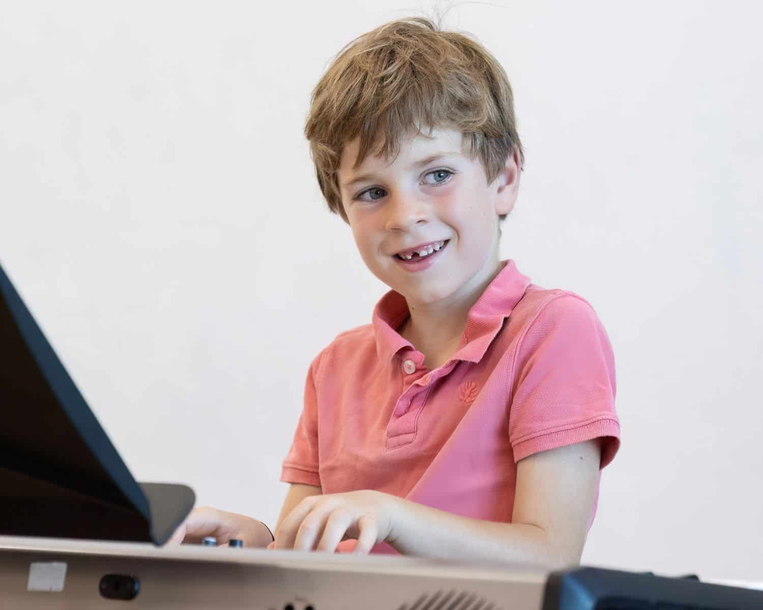 A young boy in a pink collared shirt smiles whilst playing an electronic keyboard, looking slightly to the side—one of the promising new TEMPO Ambassadors sharing his love for music.