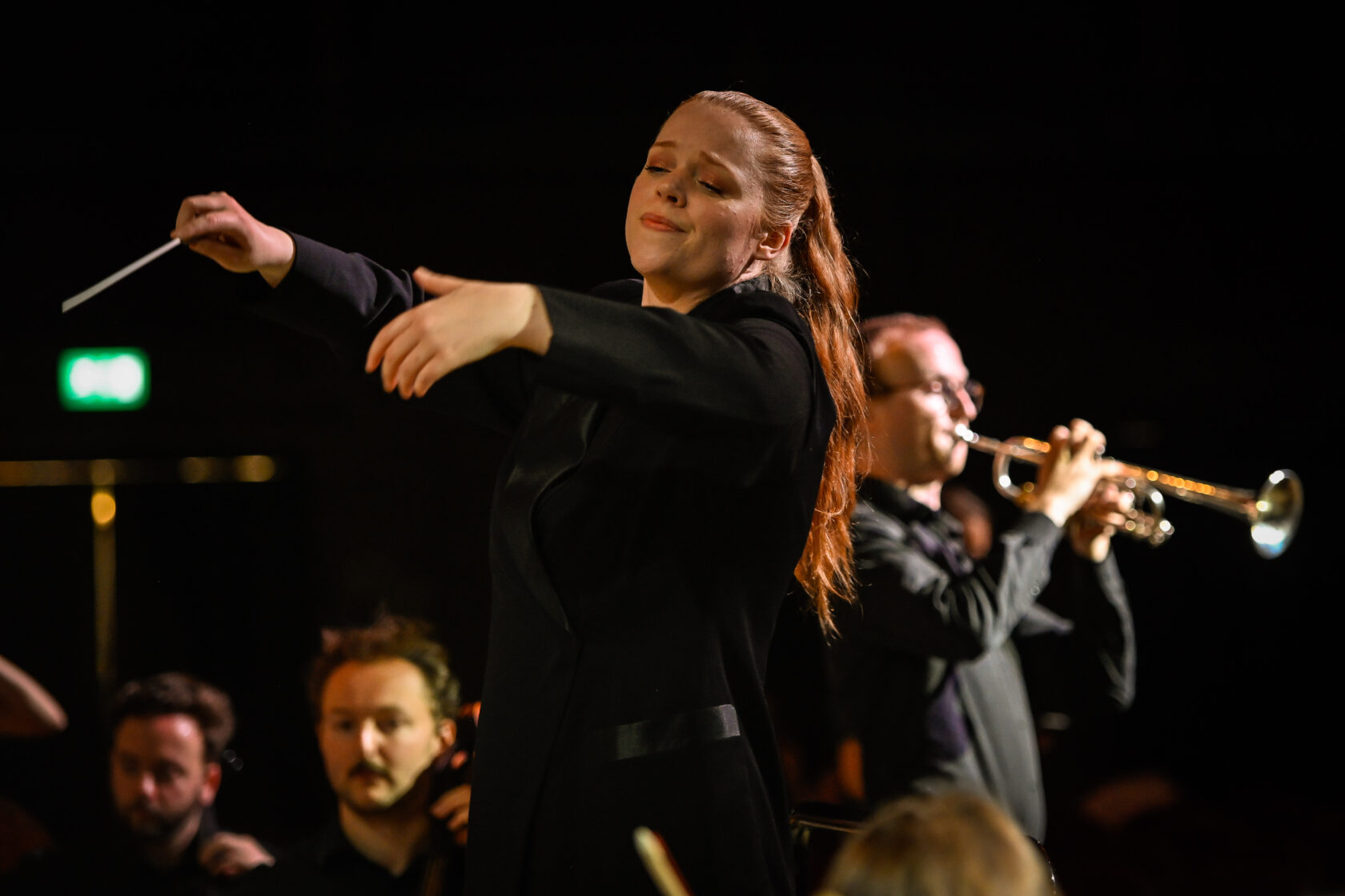 A conductor passionately leads an orchestra, eyes closed, whilst a musician in the background plays the trumpet. The scene is warmly lit, highlighting the expressive atmosphere.
