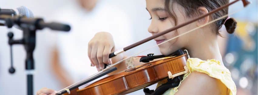 A young girl in a yellow dress, one of the New TEMPO Ambassadors, plays the violin with focus, standing near a microphone. The background is softly blurred.