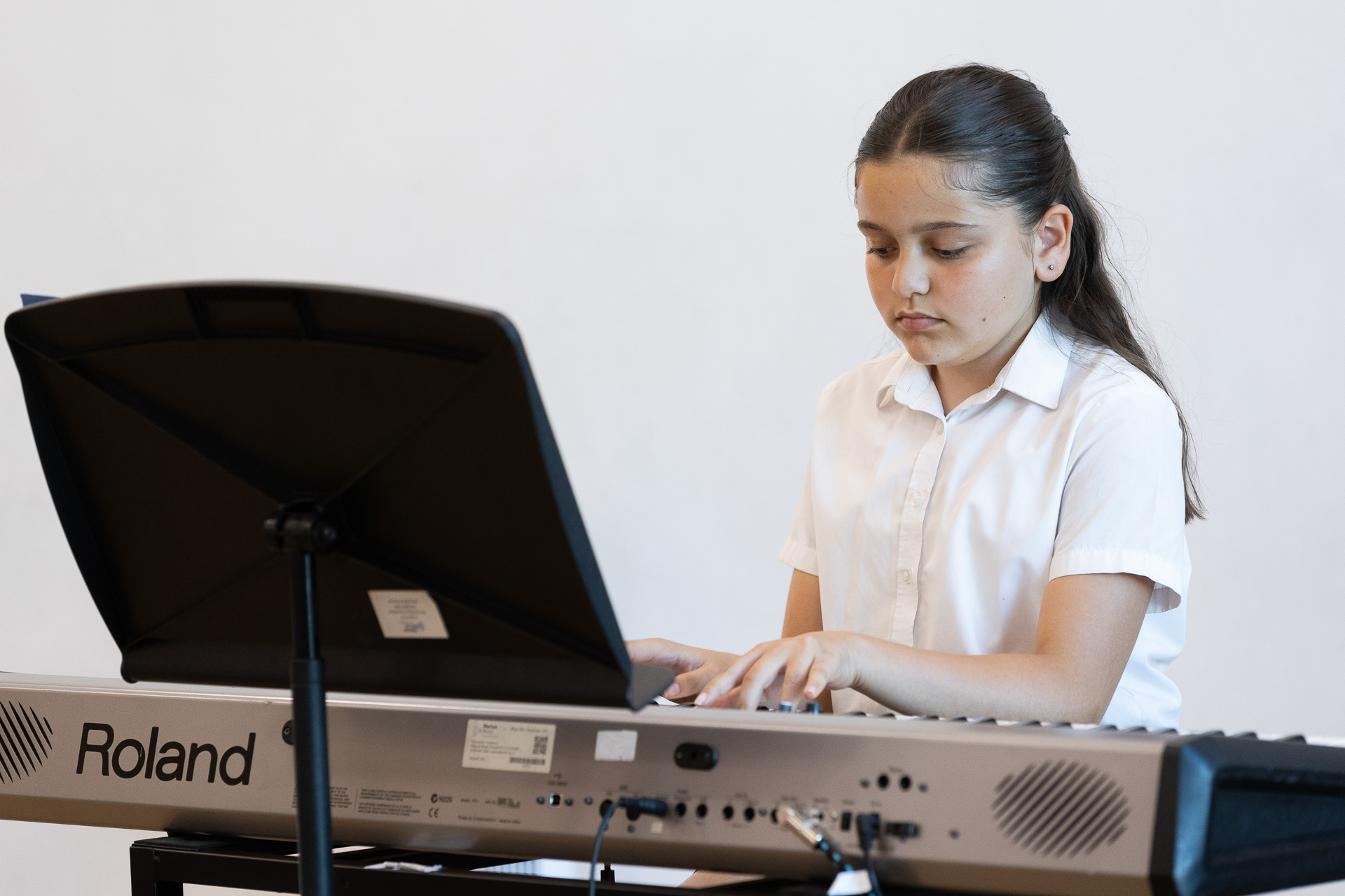 MMF.Spotlight.Summer26.054.web A young girl wearing a white shirt plays a Roland keyboard, focused on her music with a music stand in front of her against a plain light background,.