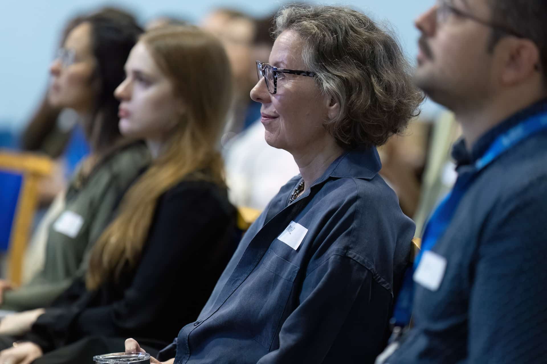 MMFTutorConference2026.03.web A group of people, including a woman with glasses, sit in a row attentively listening at an event or conference. Wearing name badges and casual business attire, the attendees include the new TEMPO Ambassadors as they engage with the presentation.