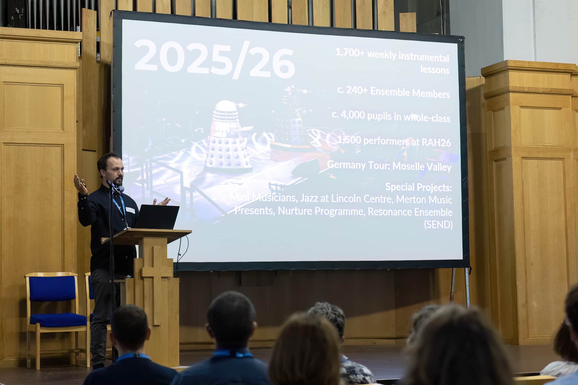 MMFTutorConference2026.05.web A man speaks at a lectern in front of an audience, presenting slides with 2025/26 music programme statistics and special projects, including tours, ensembles, and the introduction of the New TEMPO Ambassadors, in a wood-panelled hall.
