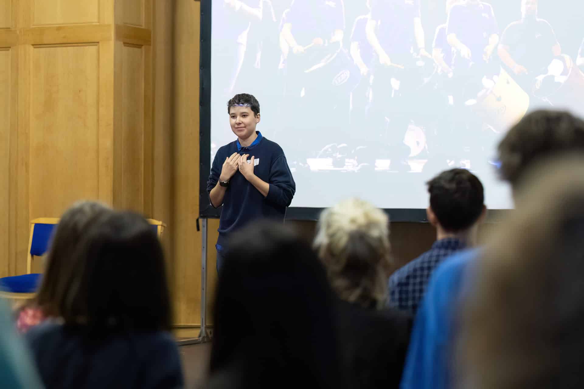 MMFTutorConference2026.09.web A new TEMPO Ambassadors representative stands in front of a projected screen, speaking to an audience in a room with wooden walls. Several people are seated or standing in the foreground, listening attentively.