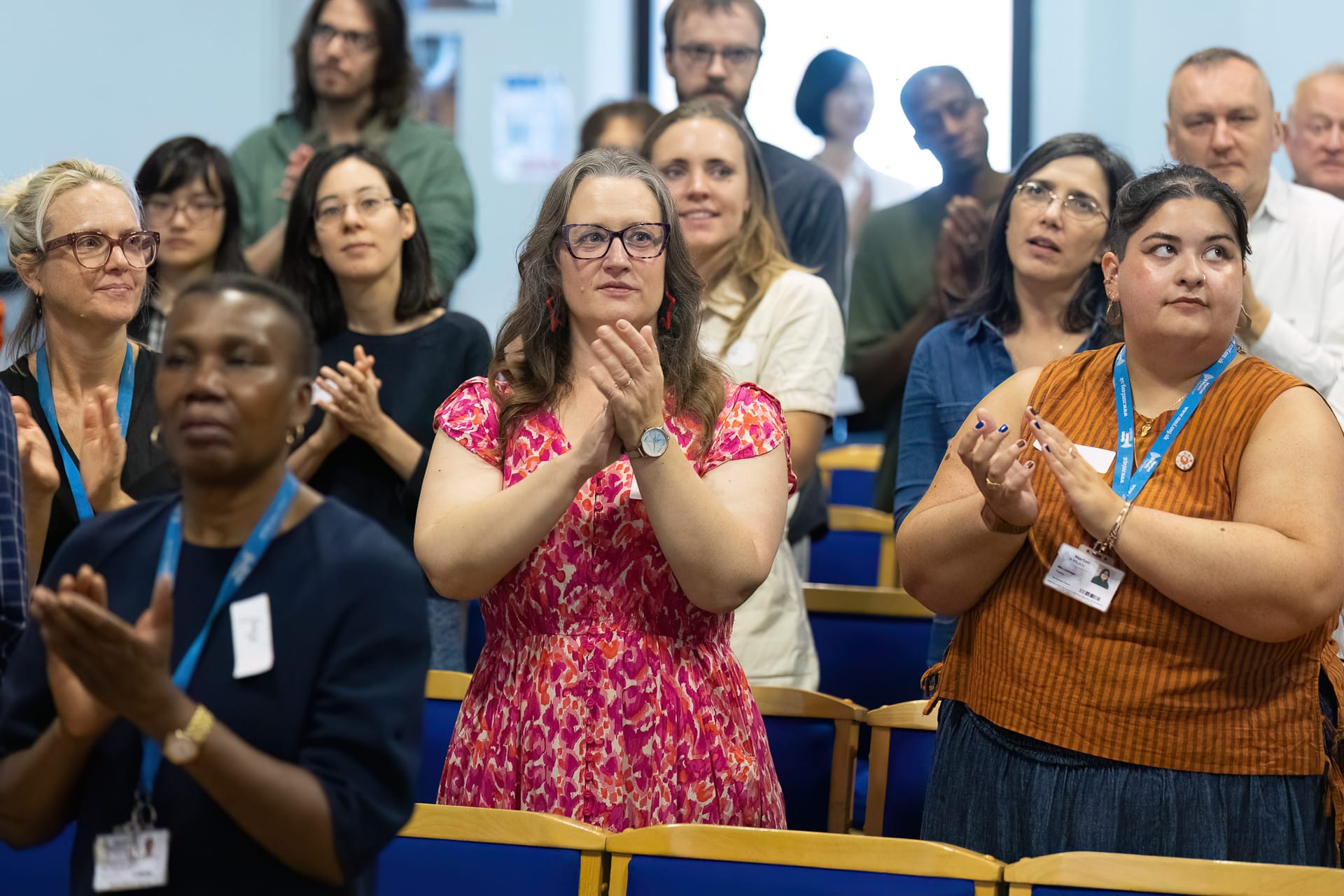 MMFTutorConference2026.13.web A diverse group of people, including the New TEMPO Ambassadors, stand and clap in a brightly lit room with blue chairs, appearing engaged and attentive—possibly at an event or presentation.