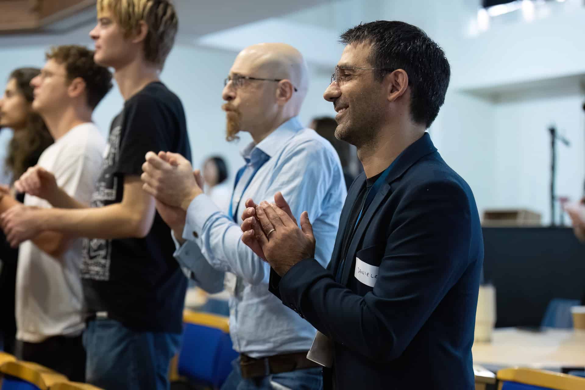 MMFTutorConference2026.19.web A group of people stands and claps at an indoor event, appearing engaged and attentive. The focus is on a smiling man in a dark jacket with a name badge, celebrated as one of the new TEMPO Ambassadors, with others clapping beside him.