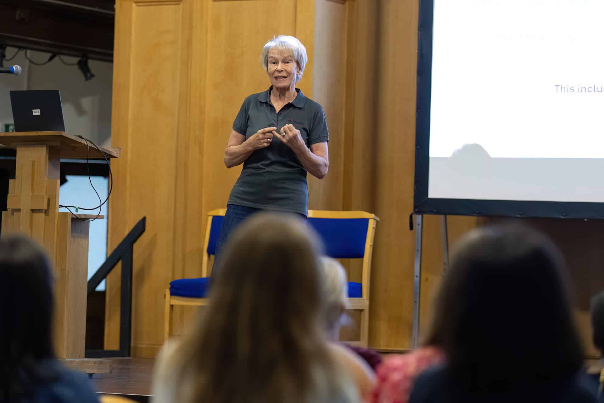 MMFTutorConference2026.21.web An older woman stands and speaks in front of an audience in a wood-panelled room, near a projection screen displaying partial text, as she welcomes the New TEMPO Ambassadors.